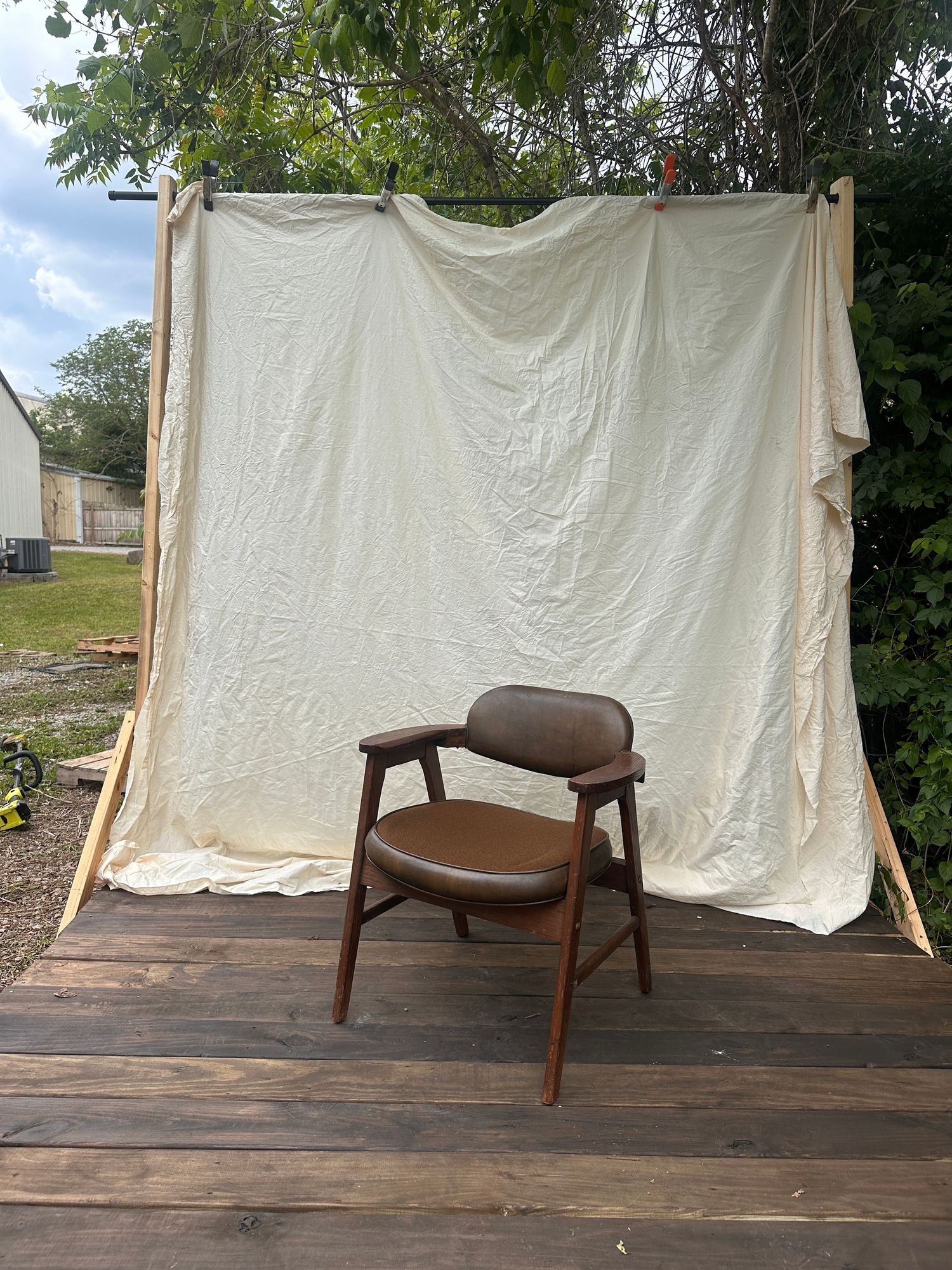 Mid-century modern brown leather armchair with walnut frame displayed on wooden deck against white fabric backdrop outdoors.