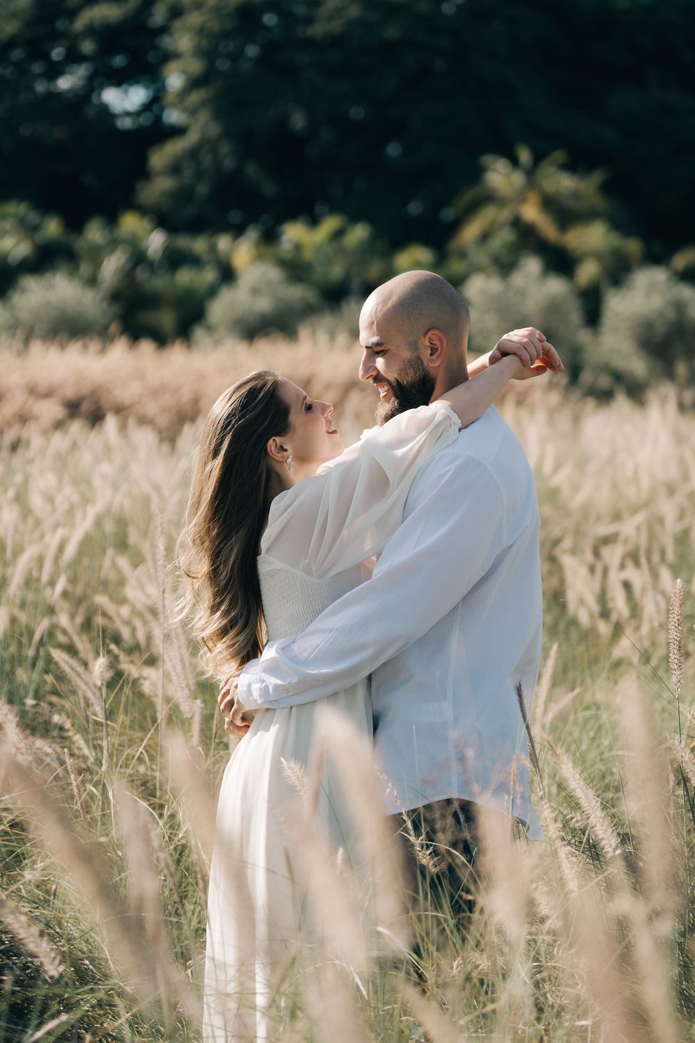 Casal se abraça em meio ao capim alto, com árvores ao fundo.
