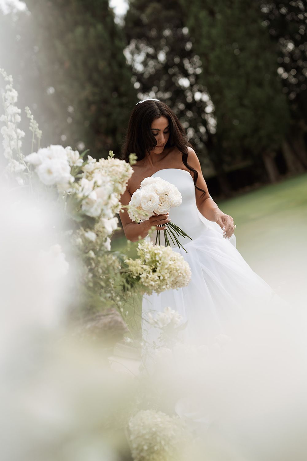 A bride in a white strapless gown holds a lush bouquet of white flowers in an outdoor garden setting.