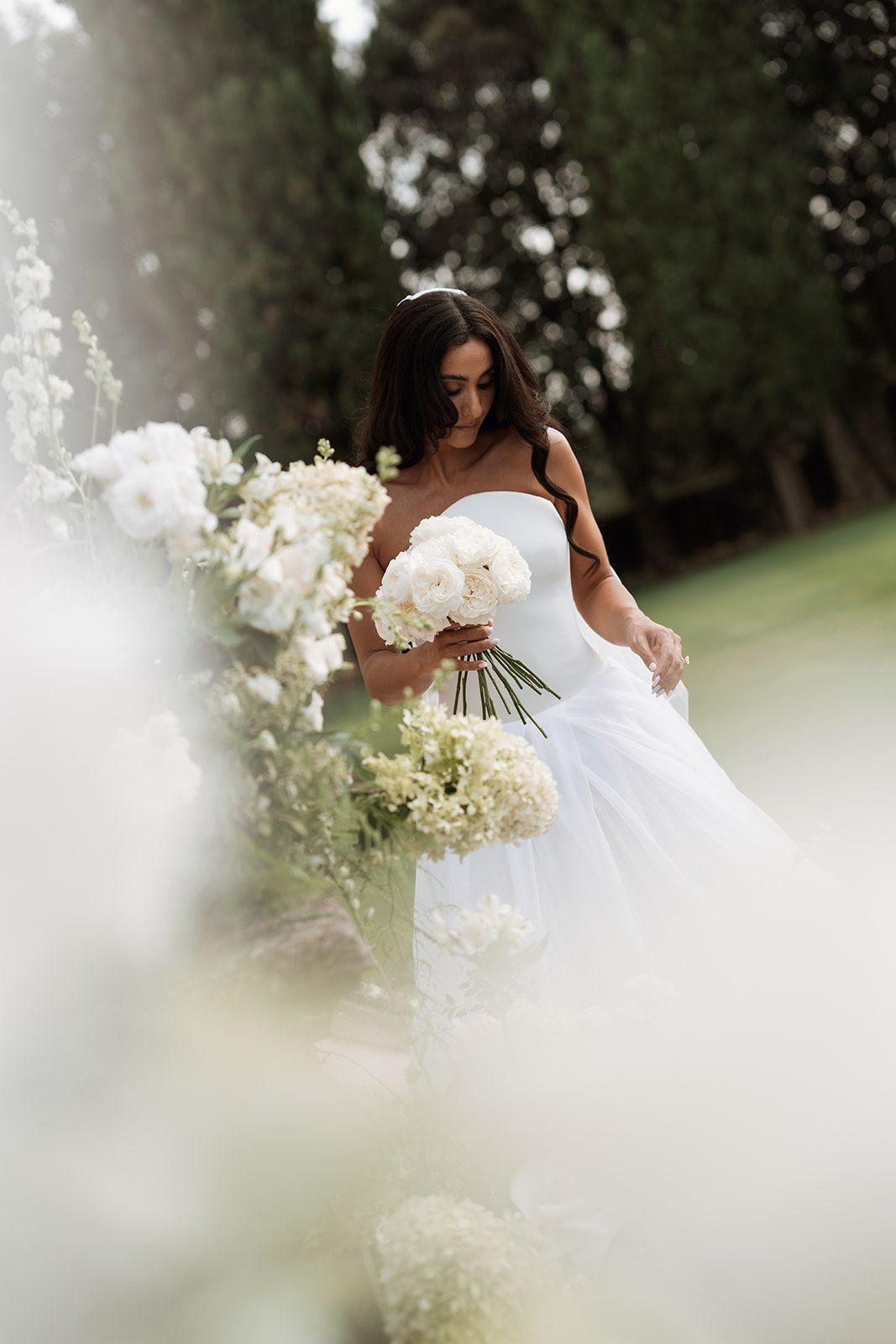 A bride in a white strapless gown holds a lush bouquet of white flowers in an outdoor garden setting.