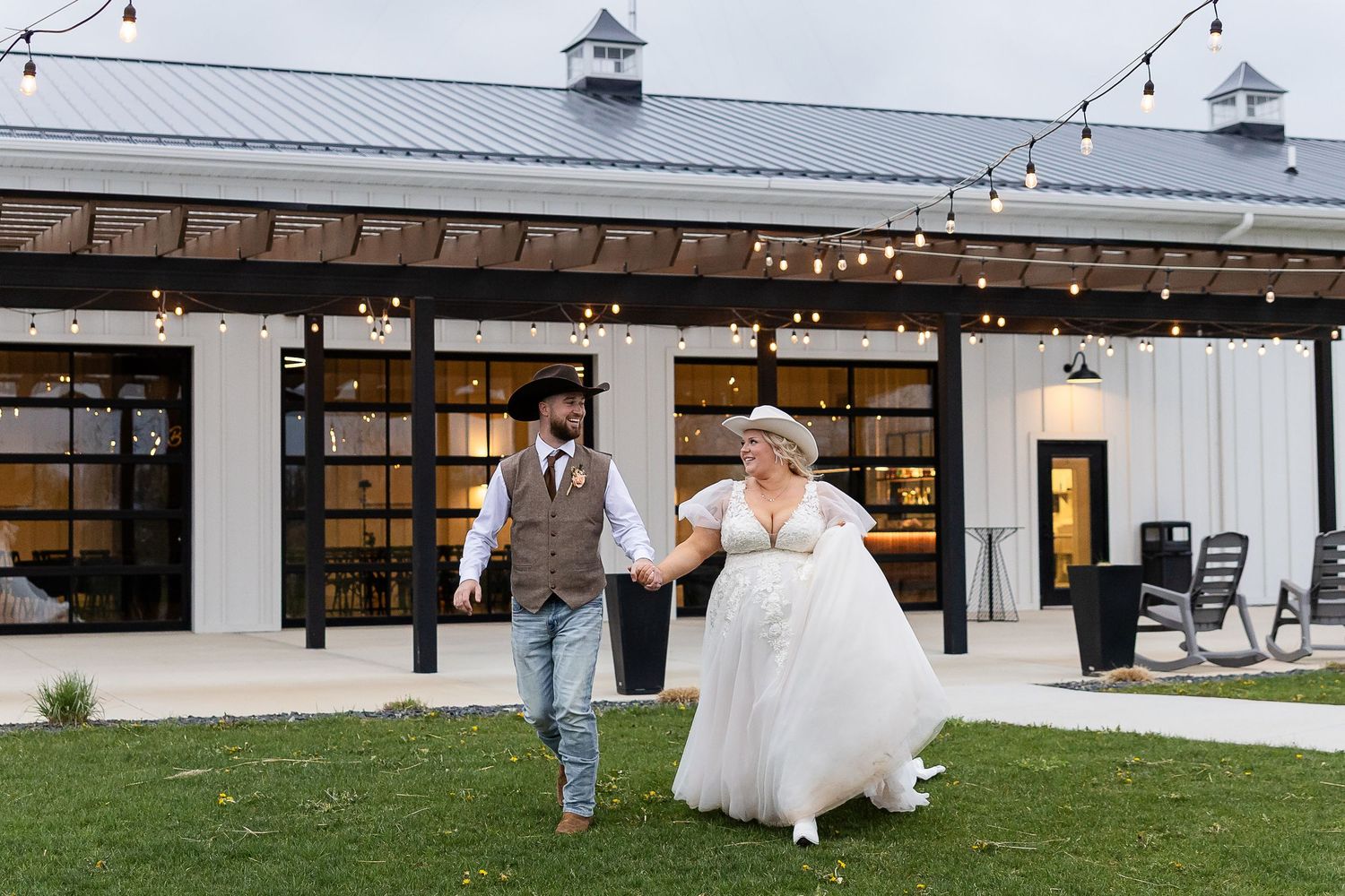 Bride and groom holding hands walking outside rustic barn wedding venue with string lights.