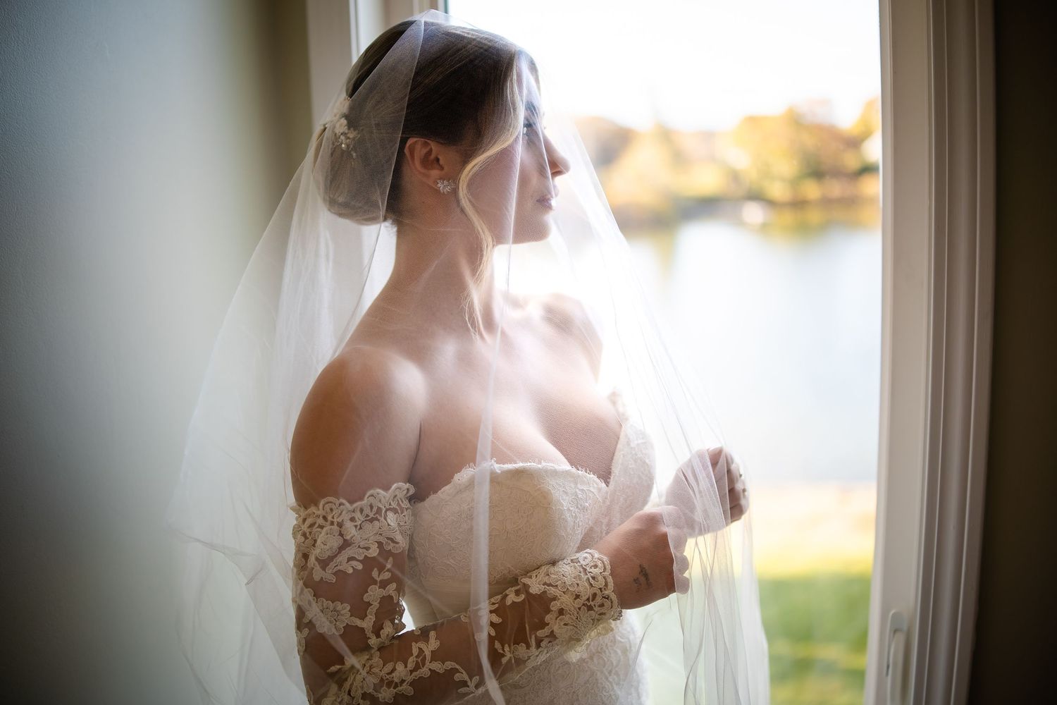 A bride in a delicate lace wedding gown and veil stands by a window overlooking a serene lake at sunset.