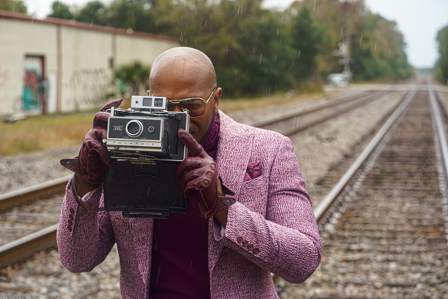 A photographer in a pink coat uses a vintage Polaroid camera while standing on railroad tracks.