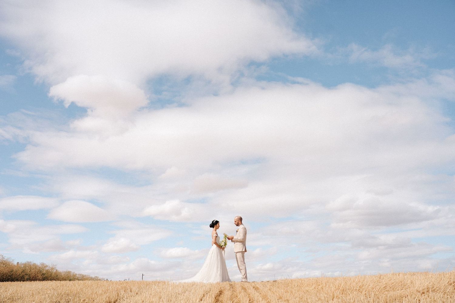A couple in white attire stands together in a golden wheat field under a bright blue sky with puffy clouds.