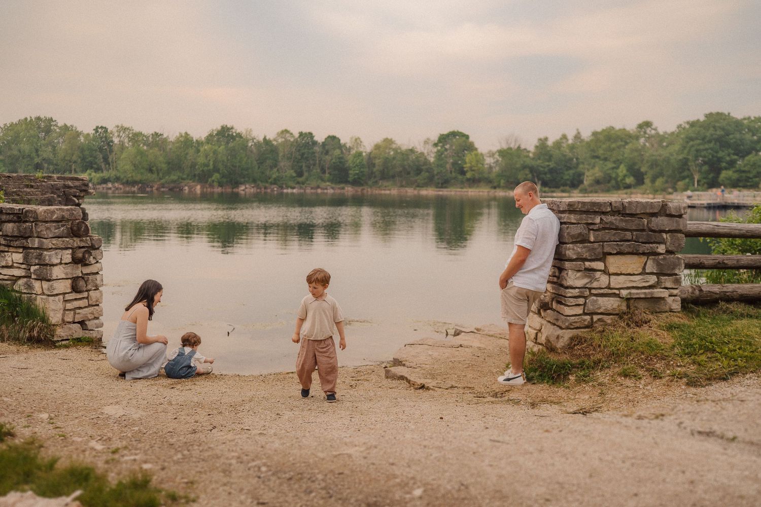 Family enjoying a peaceful afternoon by a tranquil lake, sitting on a stone wall path near the water's edge.