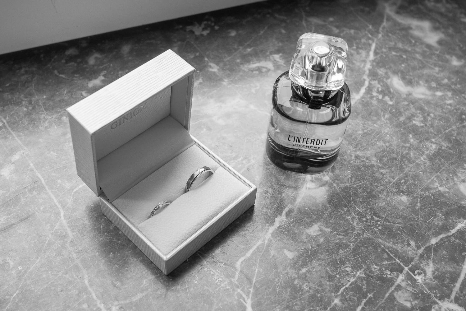 Black and white image of an engagement ring box and perfume bottle on a marble surface.