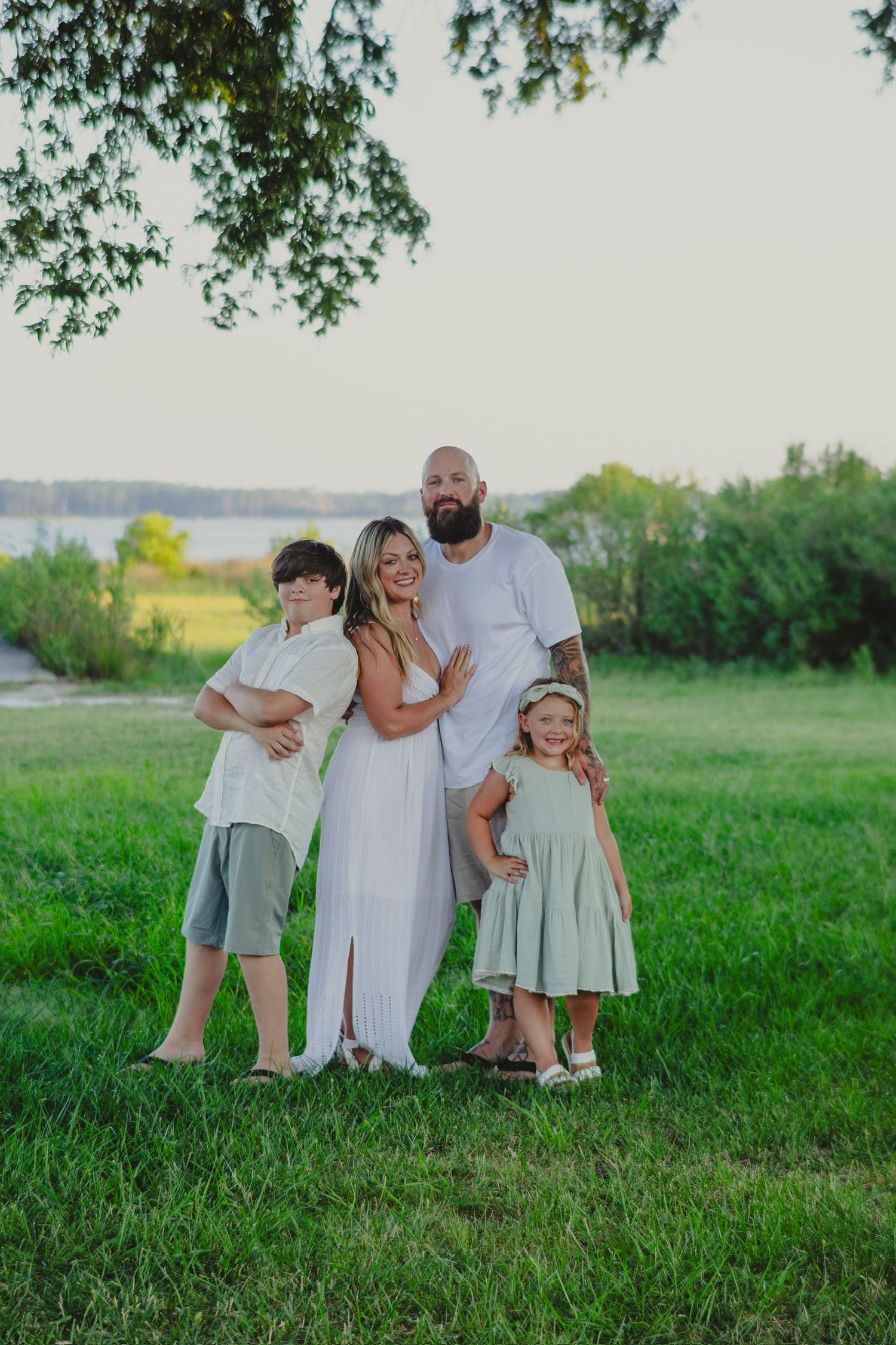 mom dad son and daughter bunched together on green grass with trees and river in the background all wearing white and sage green