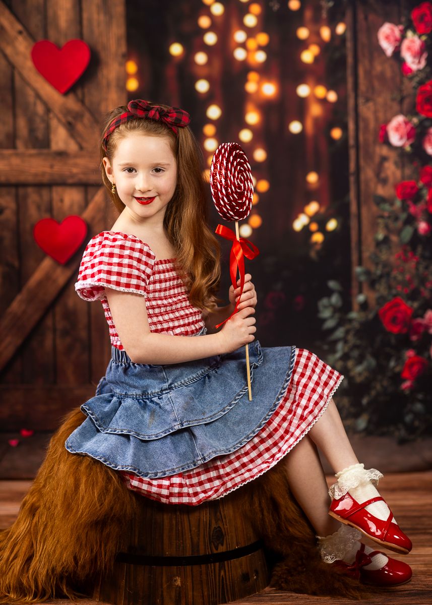Child in red gingham dress and red shoes sits on rustic wooden barrel with Valentine's Day decor and twinkle lights.