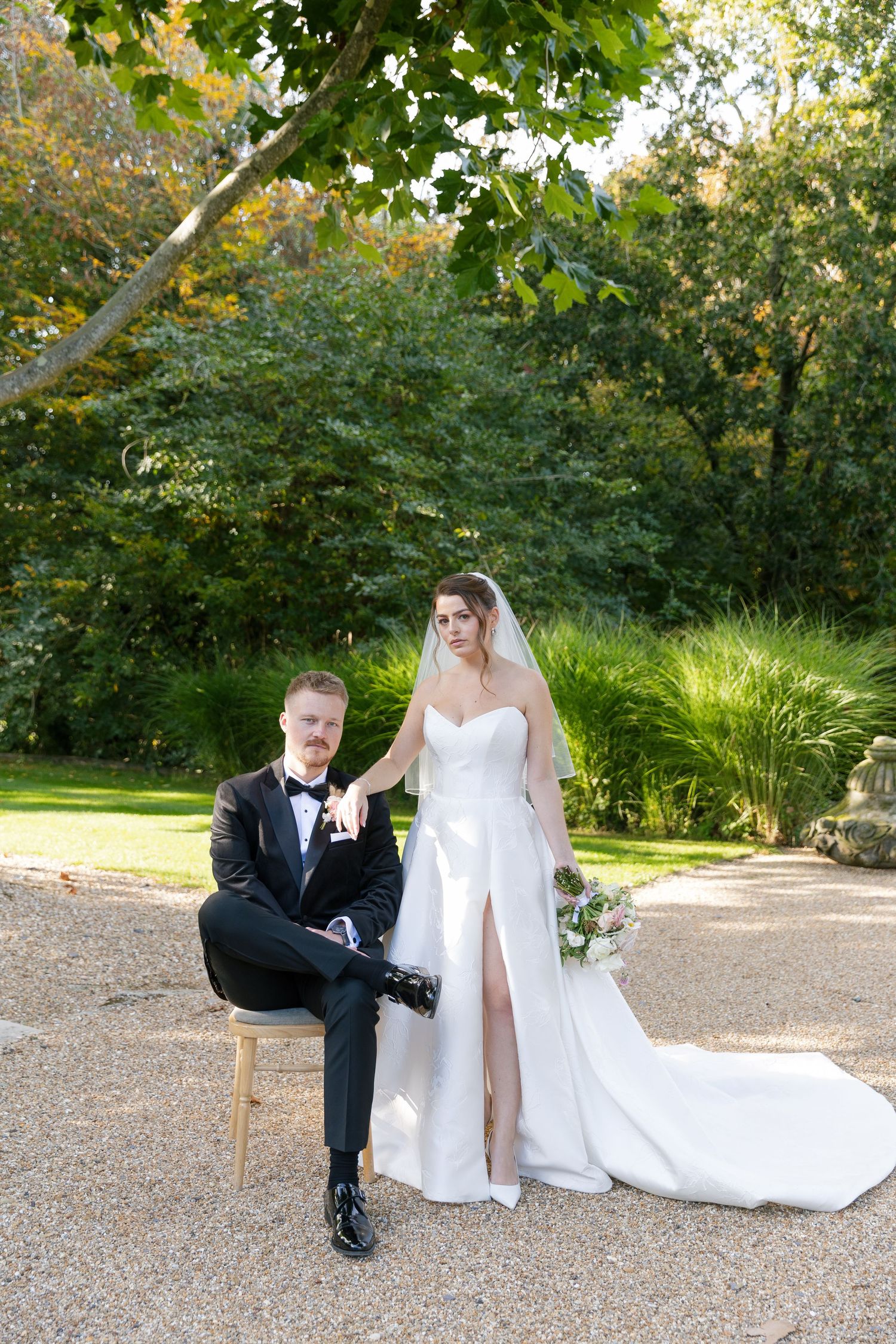 A wedding couple poses outdoors in a garden setting with lush greenery and trees in the background.
