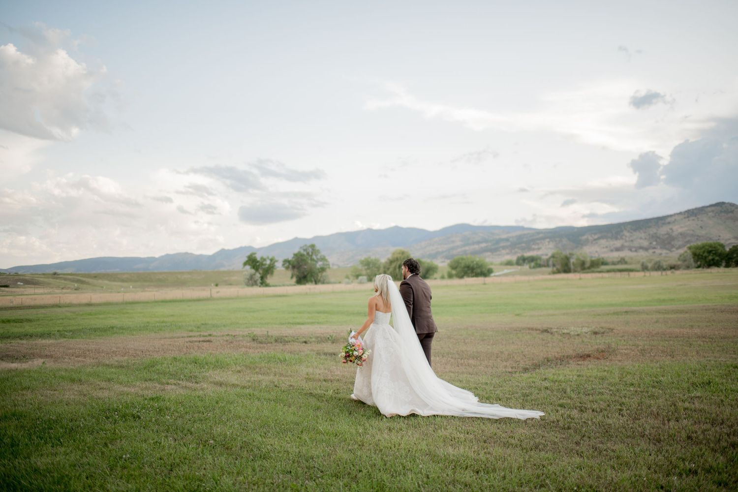 A couple in wedding attire walks through rolling green hills and meadows under a cloudy sky.