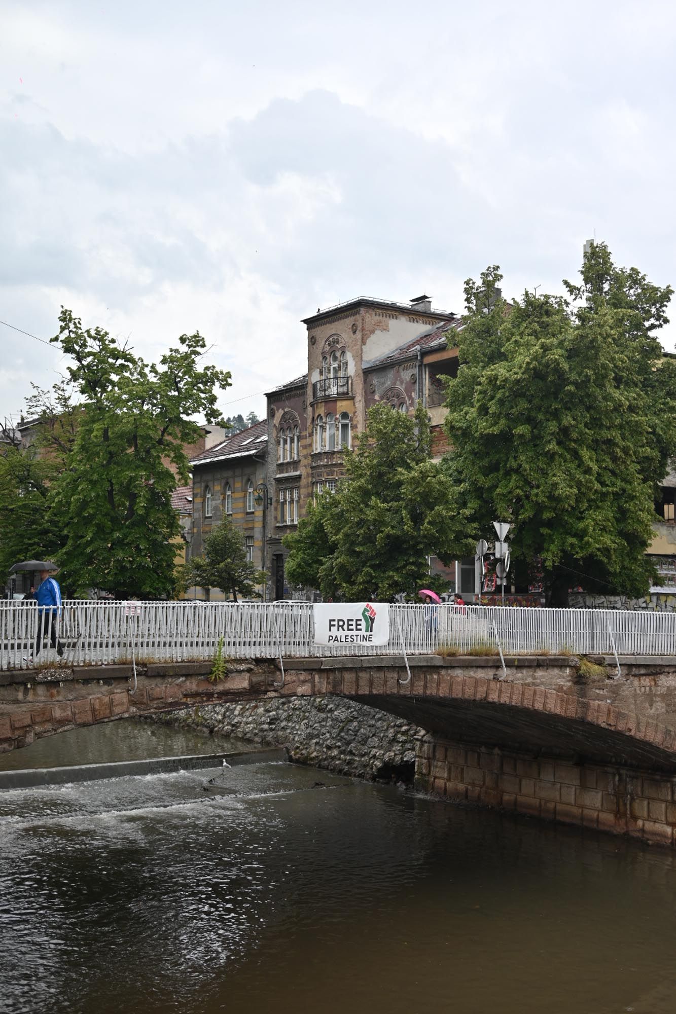 Historic stone bridge crosses a river in front of old European architecture with trees and cloudy sky in the background.