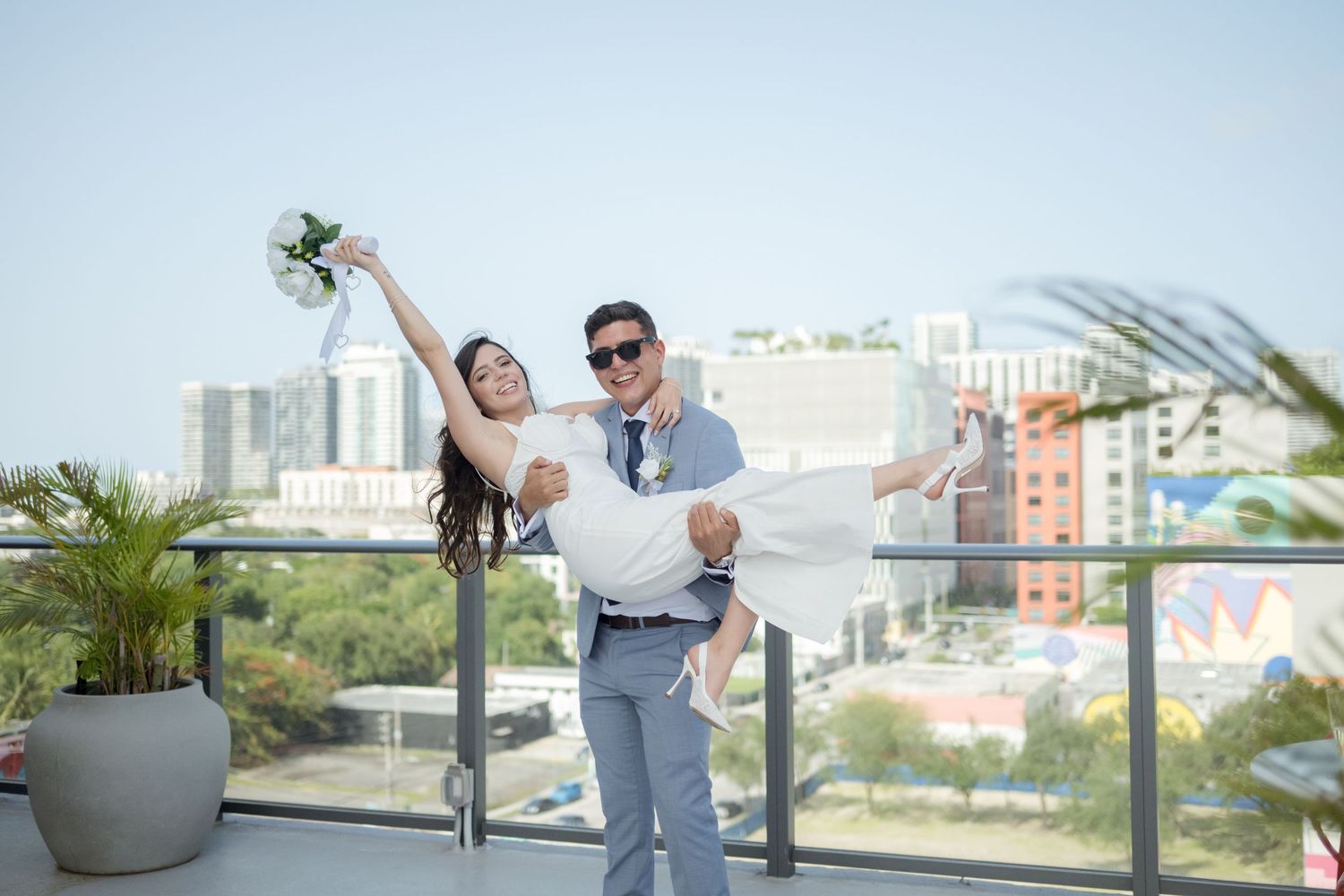 A couple in formal attire pose playfully on a rooftop terrace with a cityscape backdrop.