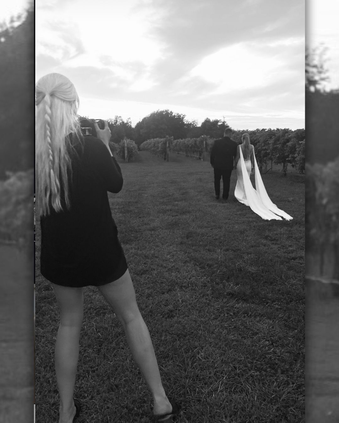 Black and white photograph of a person bending over in a grassy field with other figures visible in the distance.