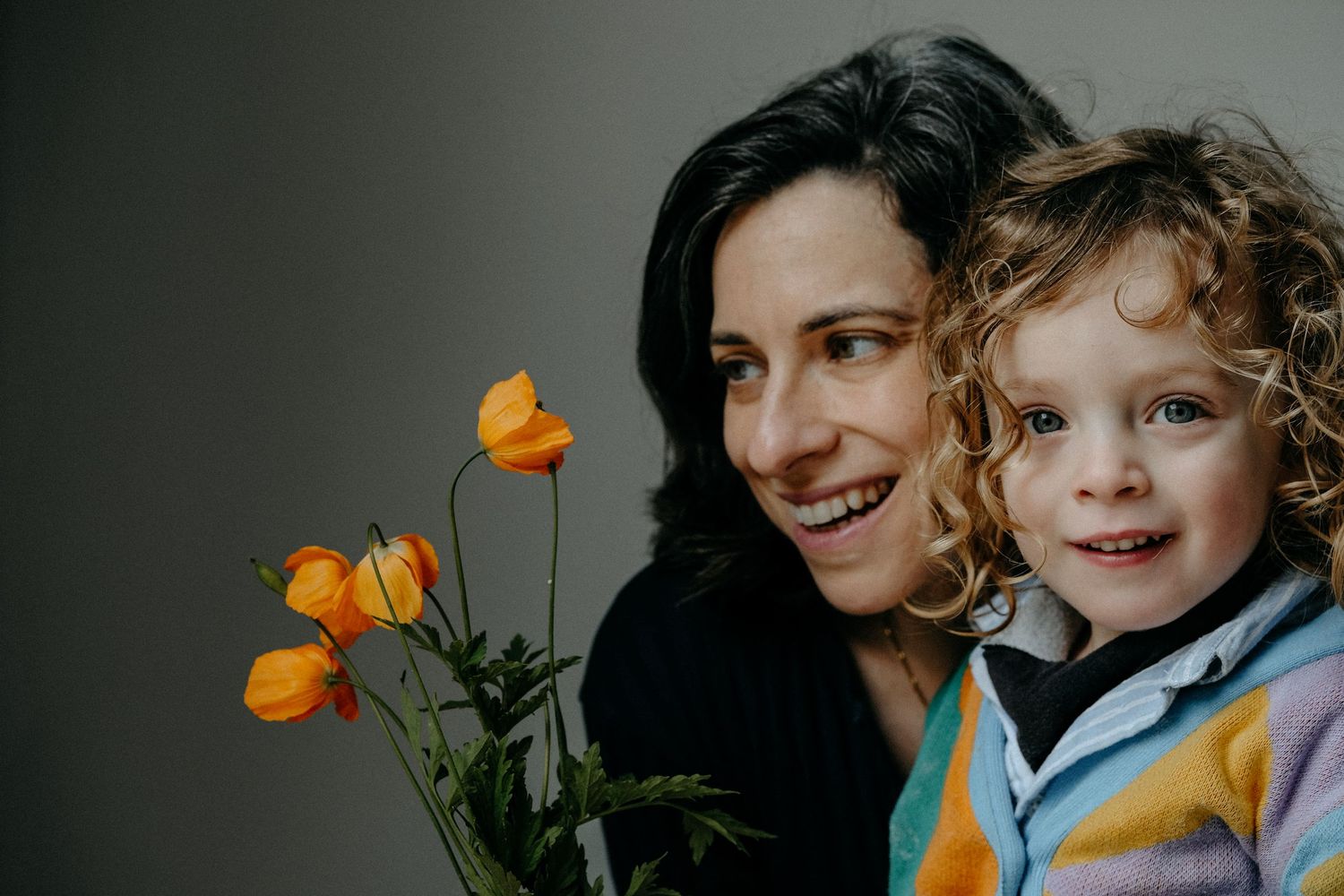 A mother and child smiling together in a warmly lit portrait, with bright orange flowers adding a pop of colour to the frame.