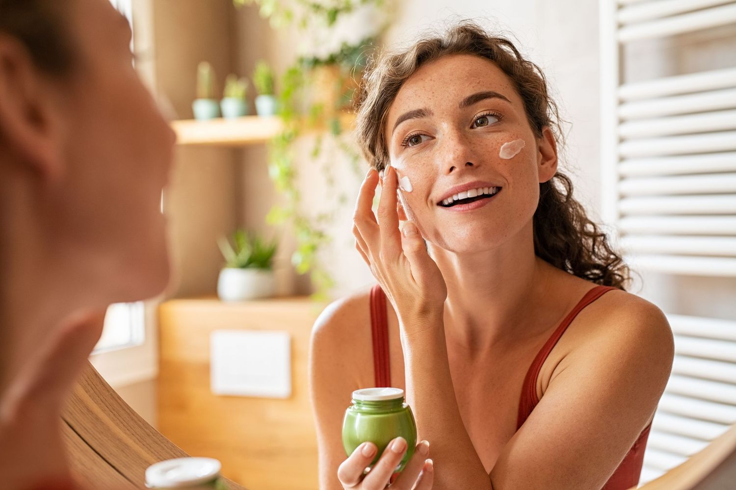 Woman applying makeup and moisturizer on her cheeks, looking in a mirror.