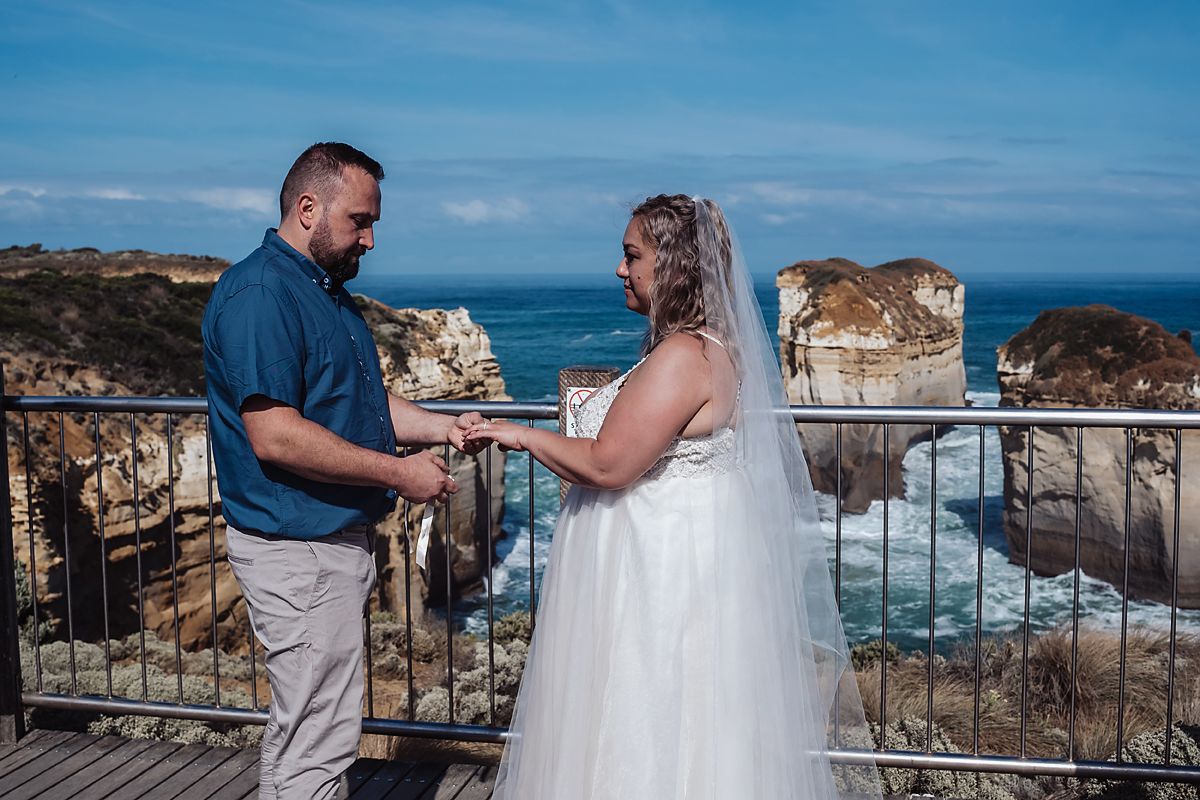Two people hold hands during a wedding ceremony at a scenic coastal overlook with limestone cliffs and ocean views.