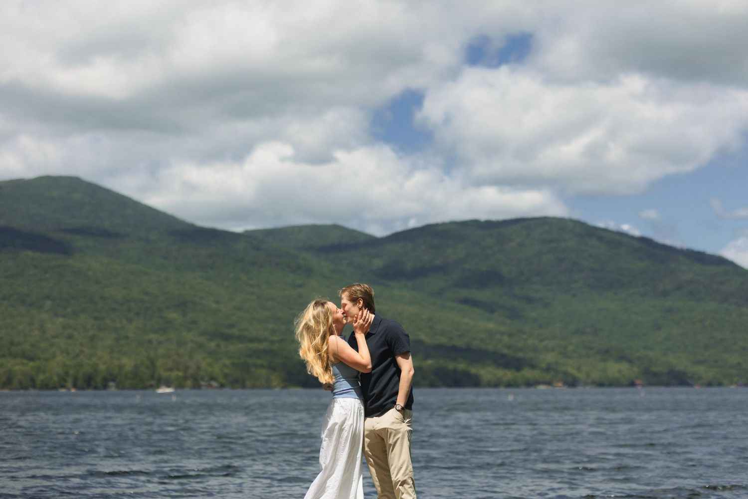 A couple embraces while standing on the shore of a lake with green mountains and cloudy skies in the background.