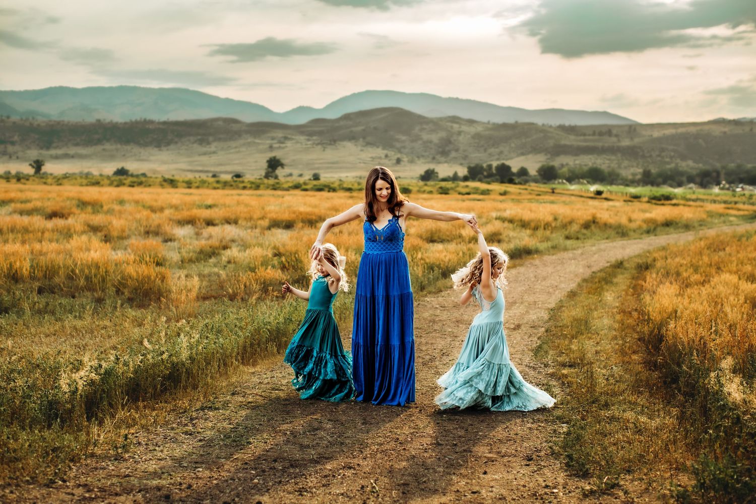 Three figures in flowing dresses stand on a dirt path through golden fields with mountains in the background.
