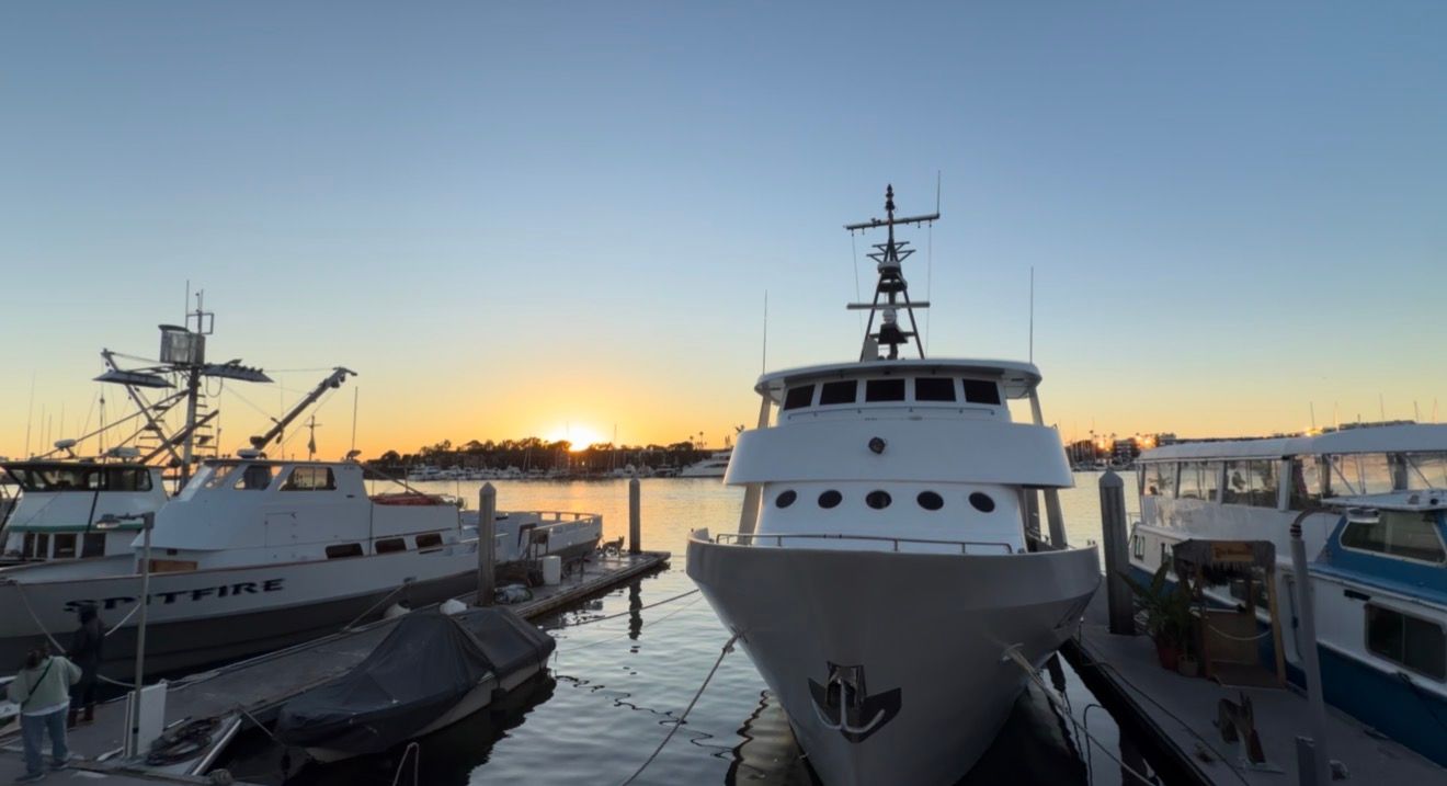 A panoramic view of boats and yachts docked at a marina during sunset with orange sky reflecting on calm waters.