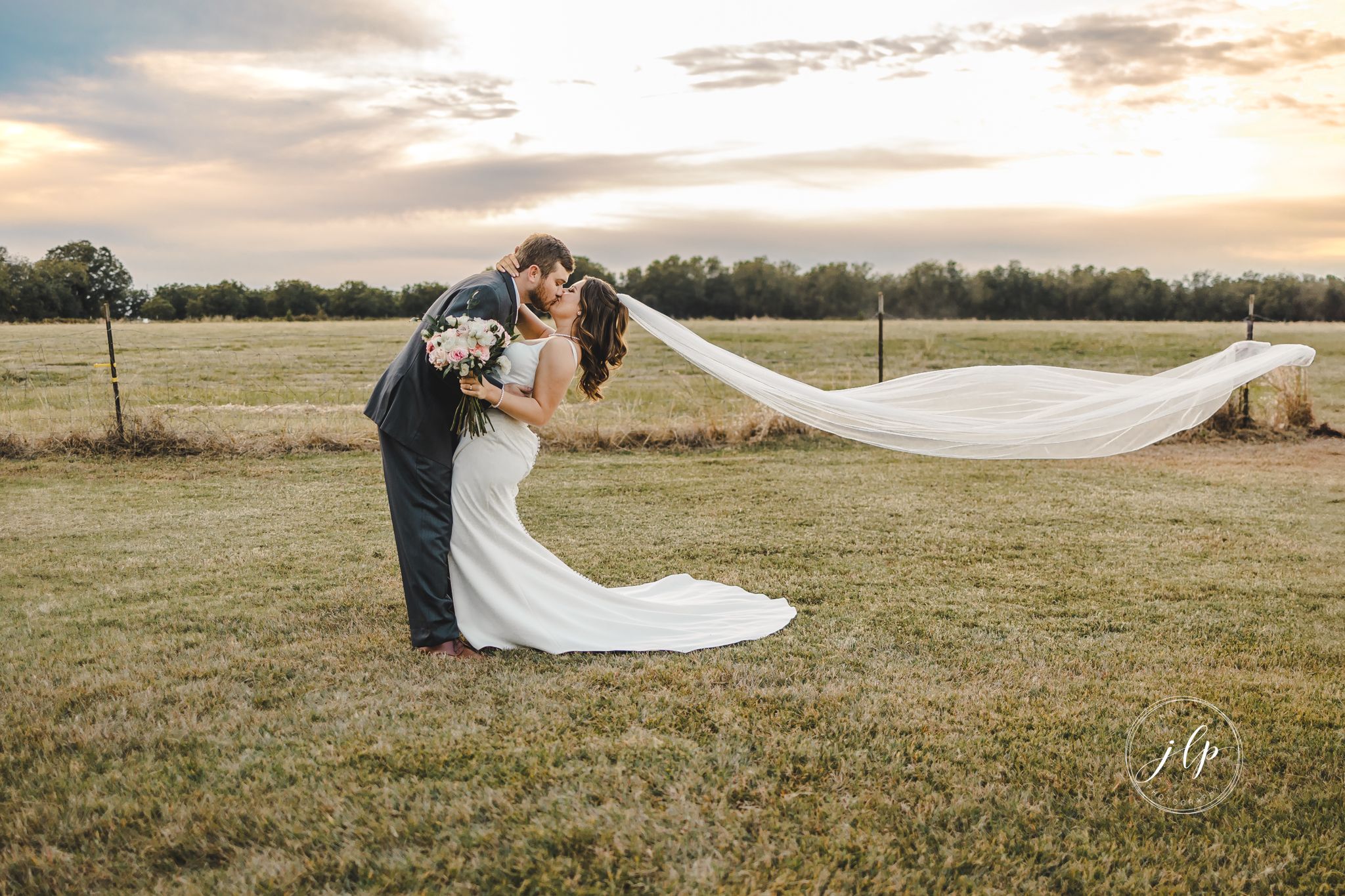 Couple embraces in a golden field during sunset with flowing wedding veil in the breeze.