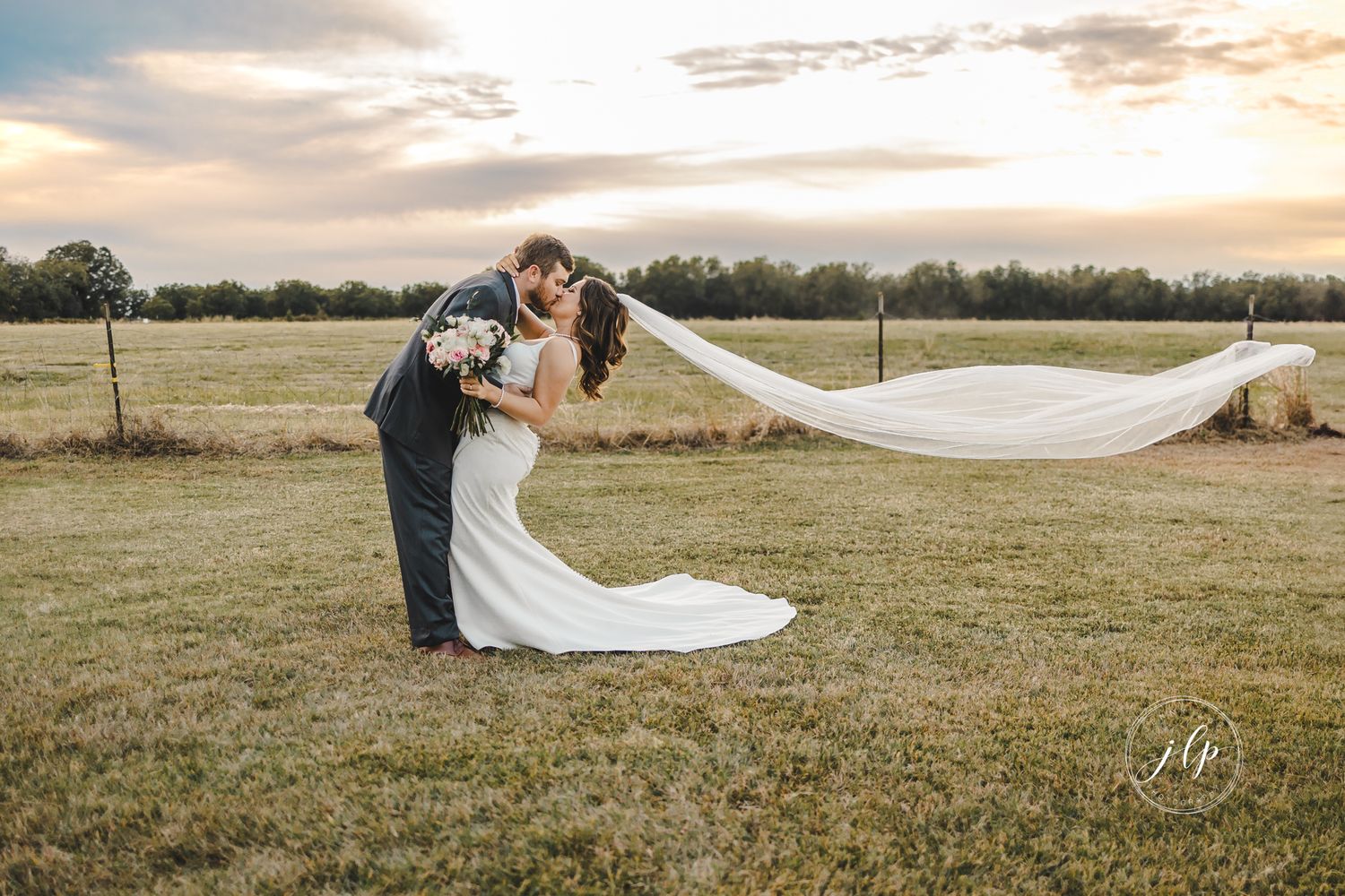 Couple embraces in a golden field during sunset with flowing wedding veil in the breeze.