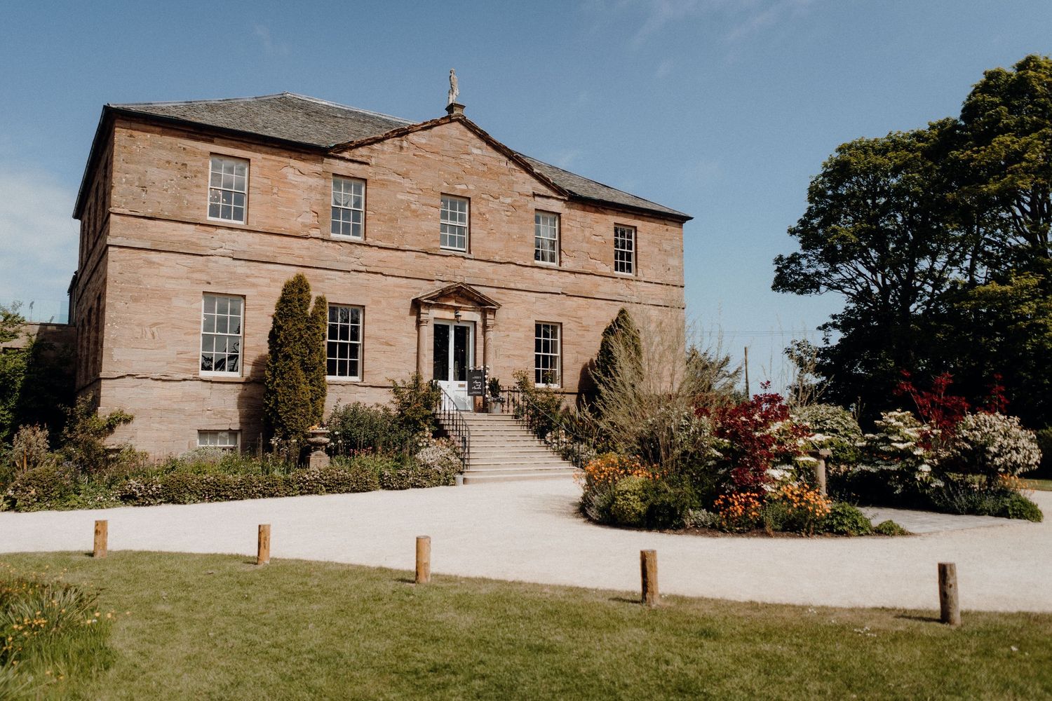 Historic stone mansion with elegant architecture set on landscaped grounds against a sunny sky.