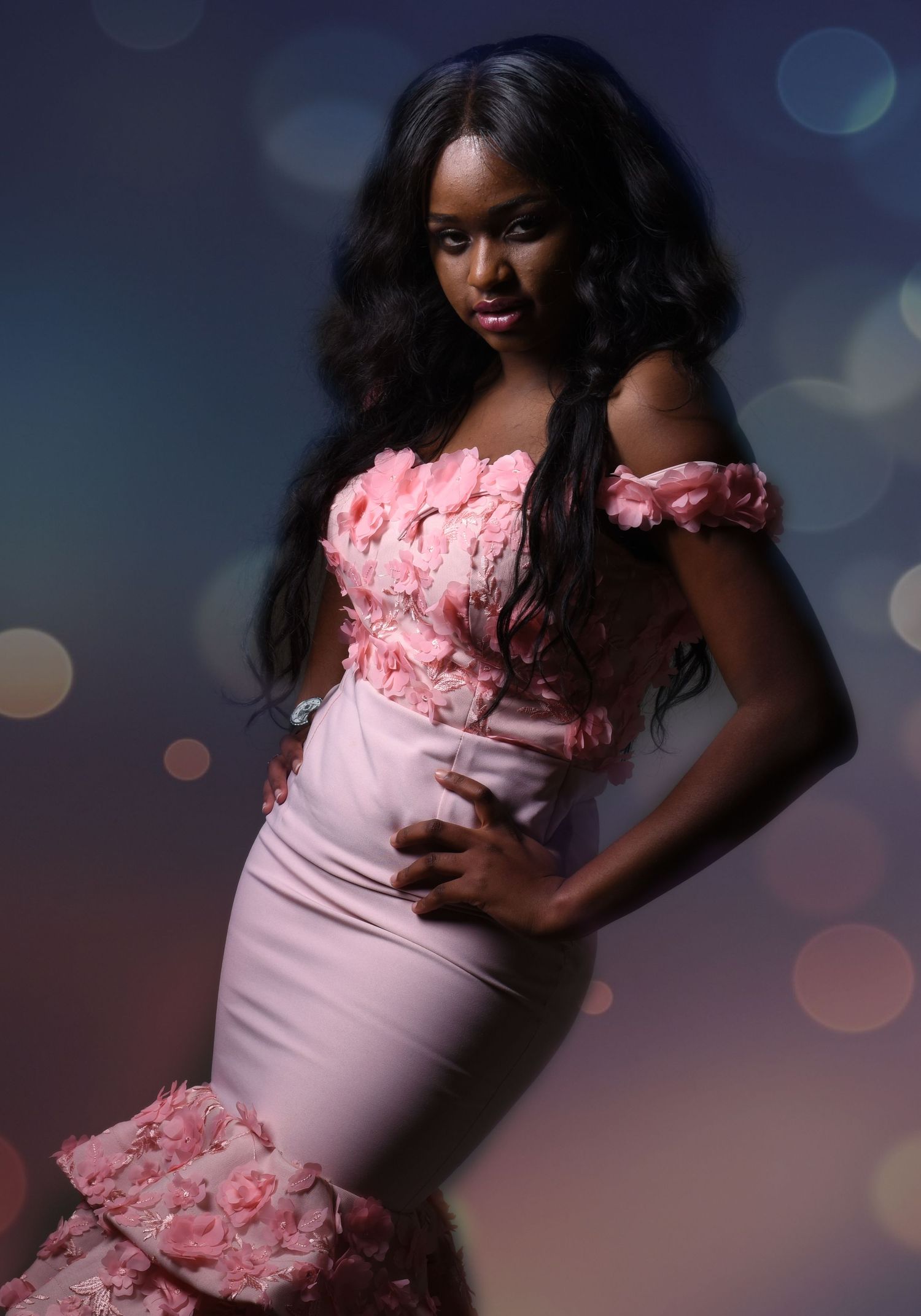 Glamorous pink mermaid-style evening gown with floral appliques and off-shoulder design photographed against blurred lights.