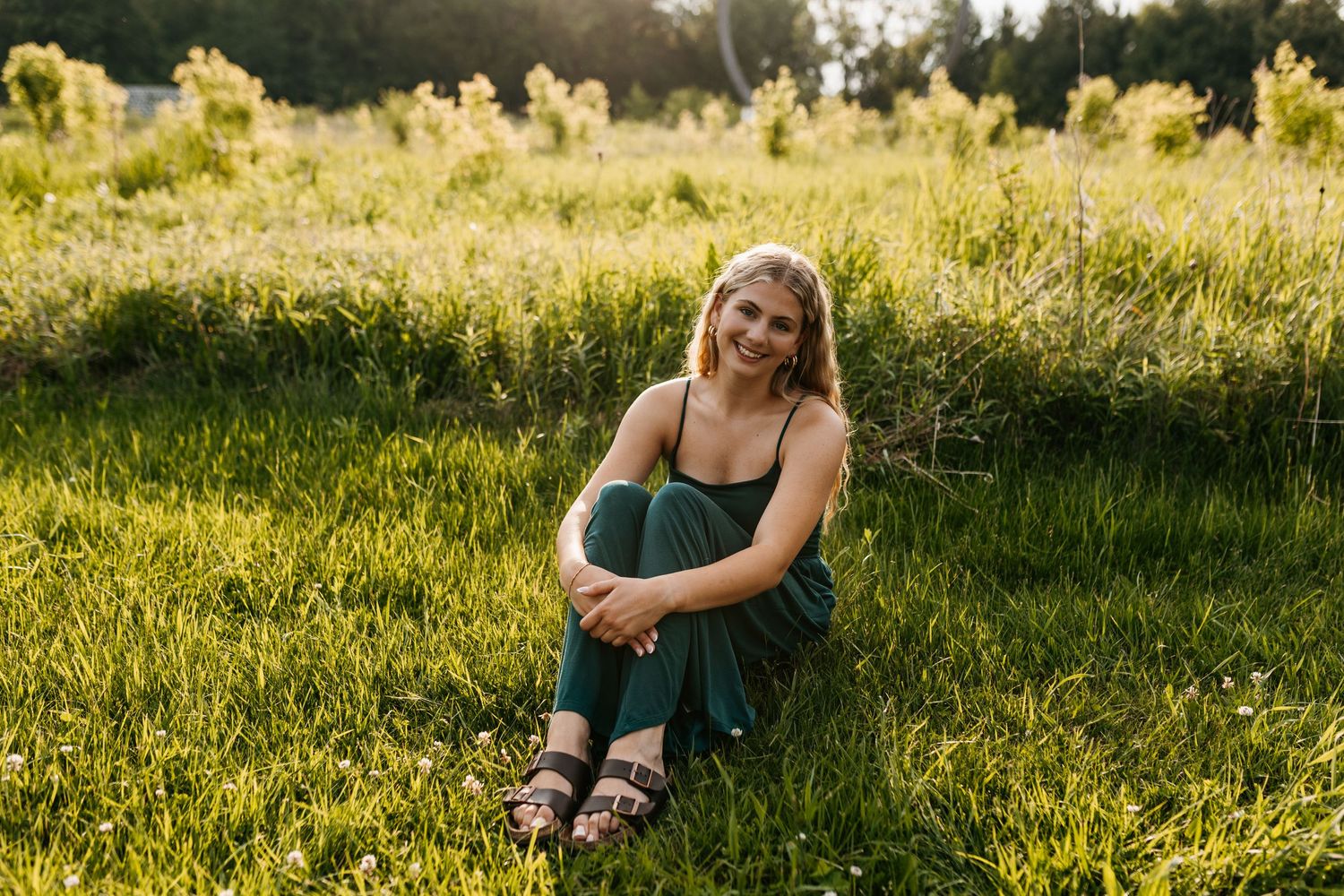 A person sits in a green field at sunset wearing a dark dress and sandals during golden hour.