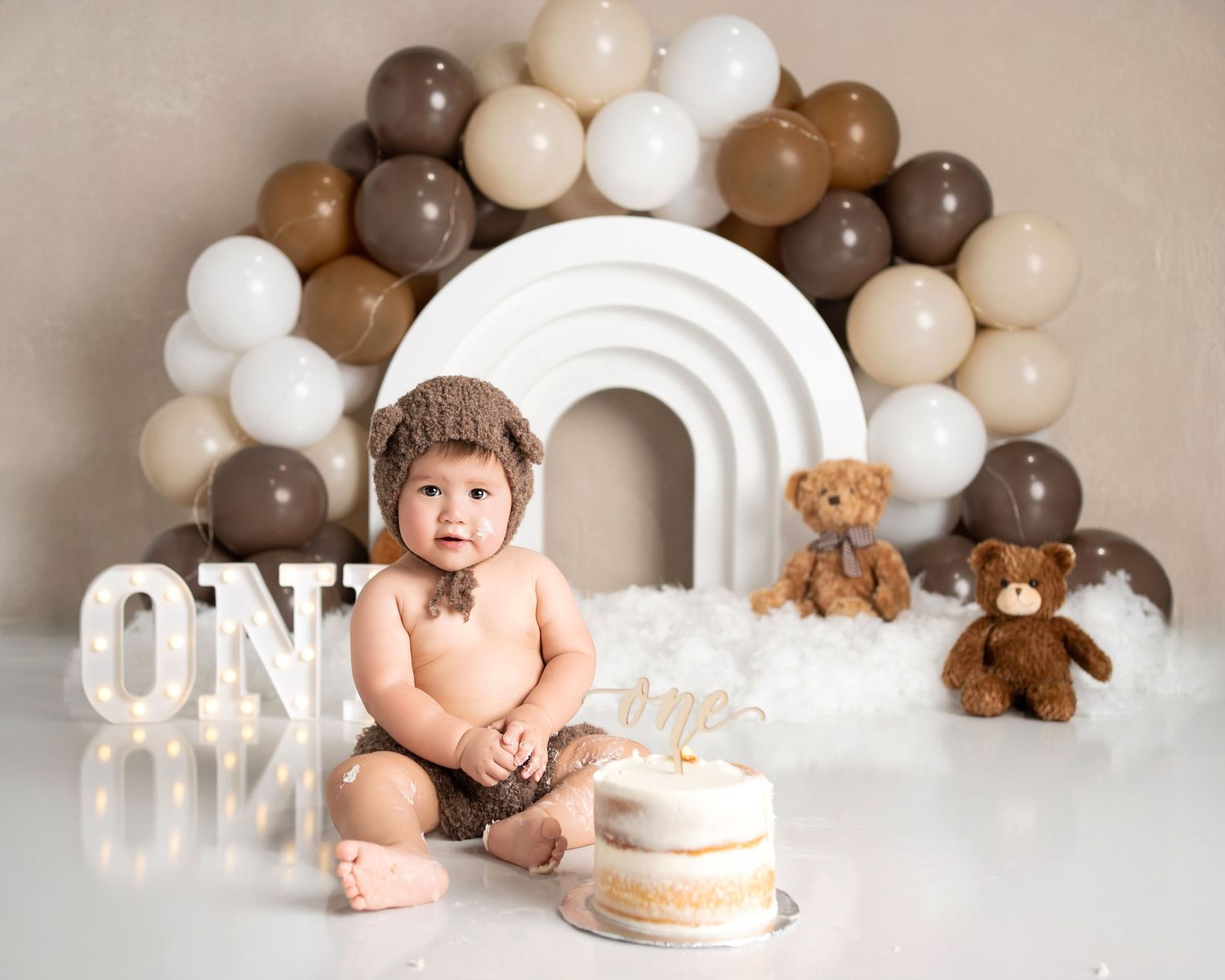 Baby in bear outfit smiling during cake smash session with brown and white balloon arch.