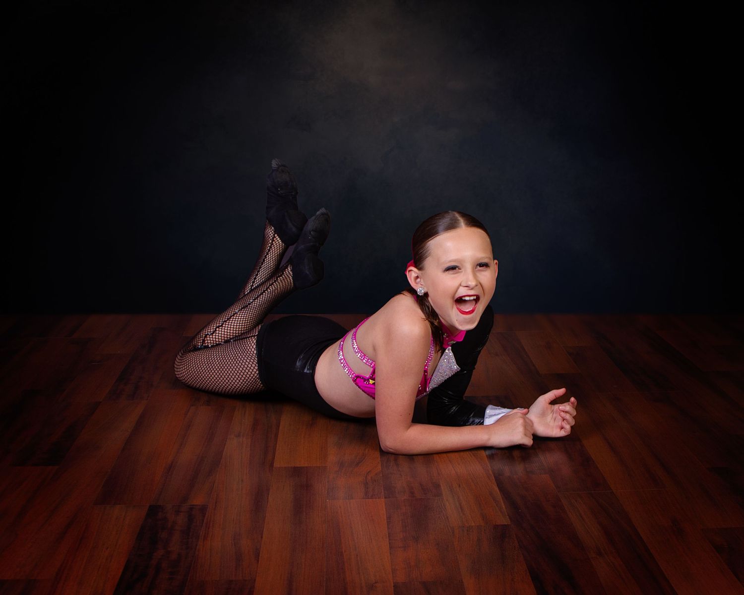 Dancer in black leotard and pink top performs dance pose laying on wooden floor in studio with dark backdrop.