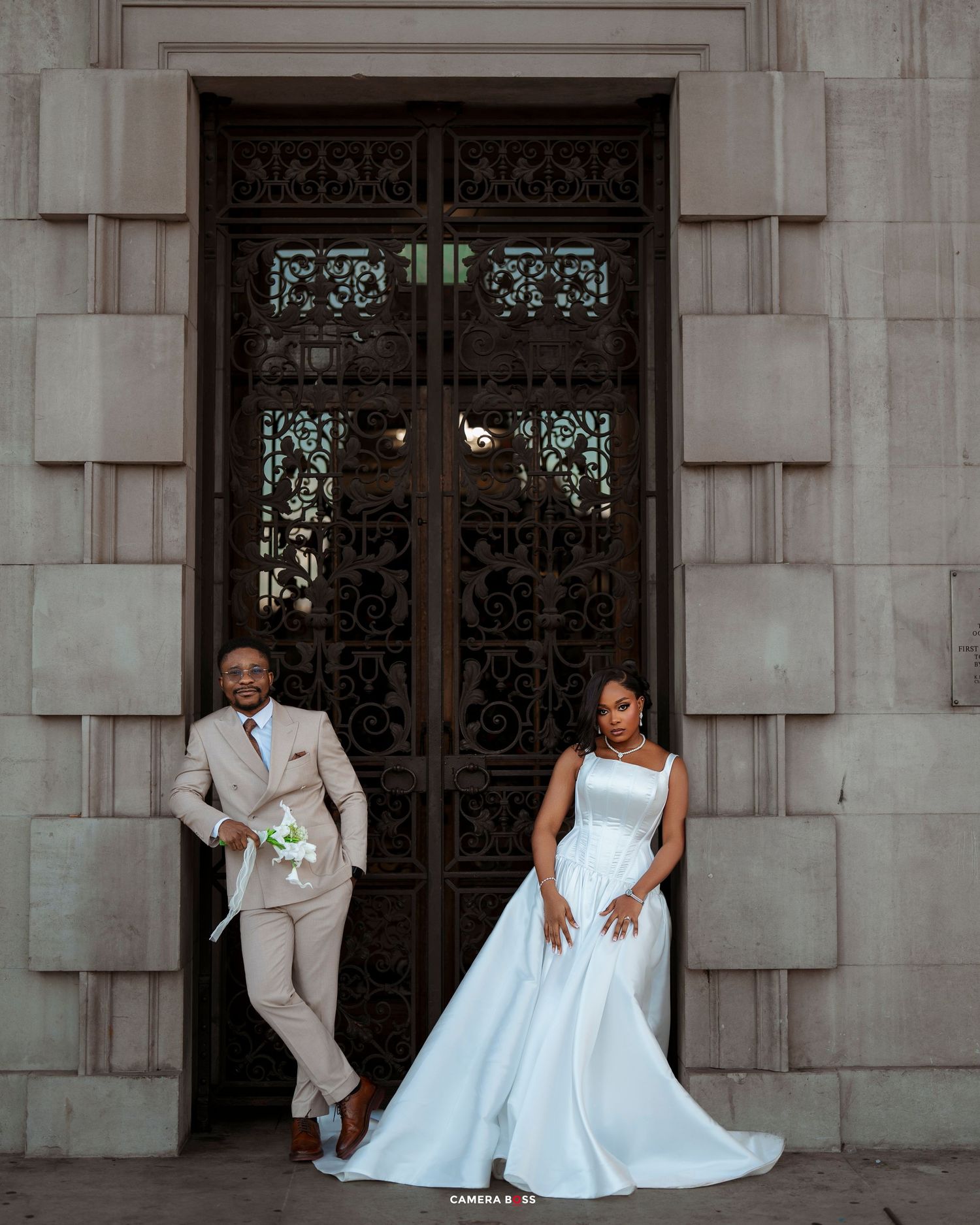 Wedding couple in formal attire poses playfully in front of ornate stone building entrance with elegant doors.