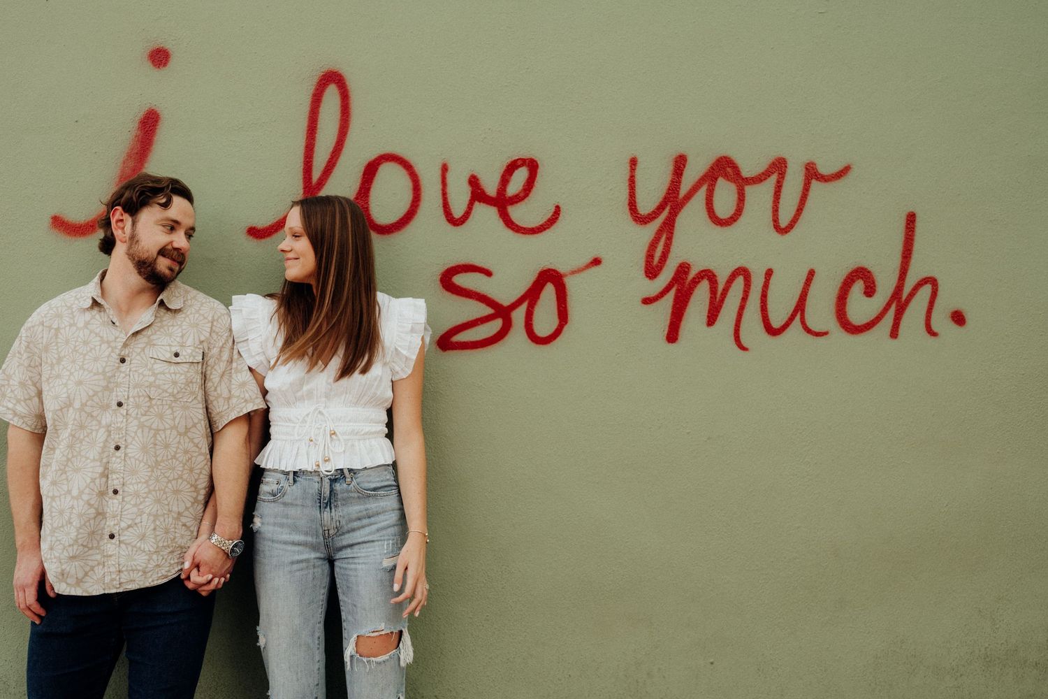 A romantic couple stands against a green wall with red text reading 'I love you so much'.