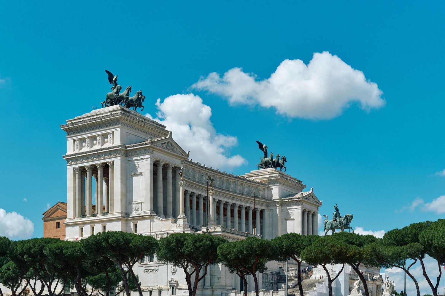 Monumental white classical building with quadriga statues on top against bright blue sky in Rome.