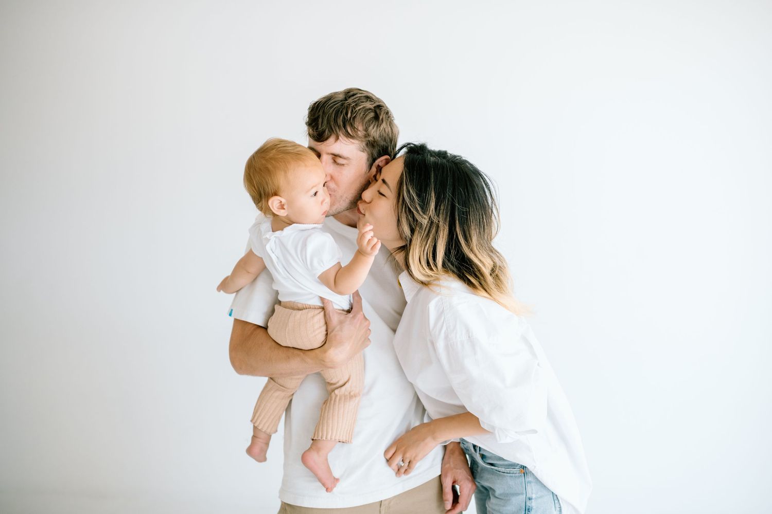 Mom leans over and kisses baby in dad's arms during family studio photoshoot