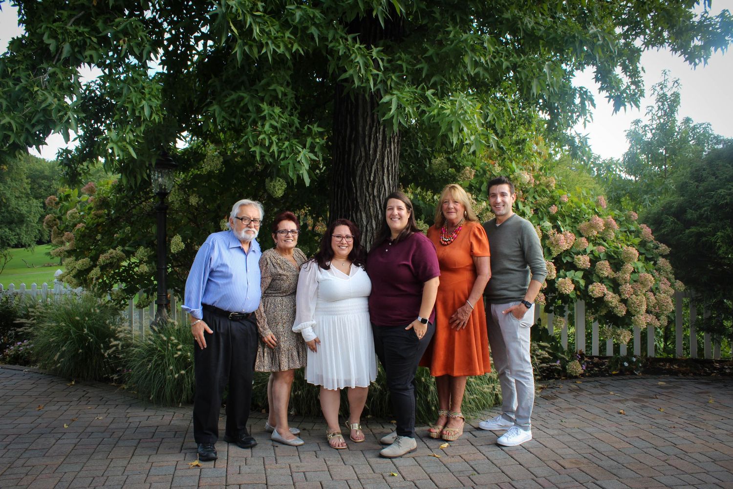 Family gathering photo on brick patio with flowering bushes and trees in background.