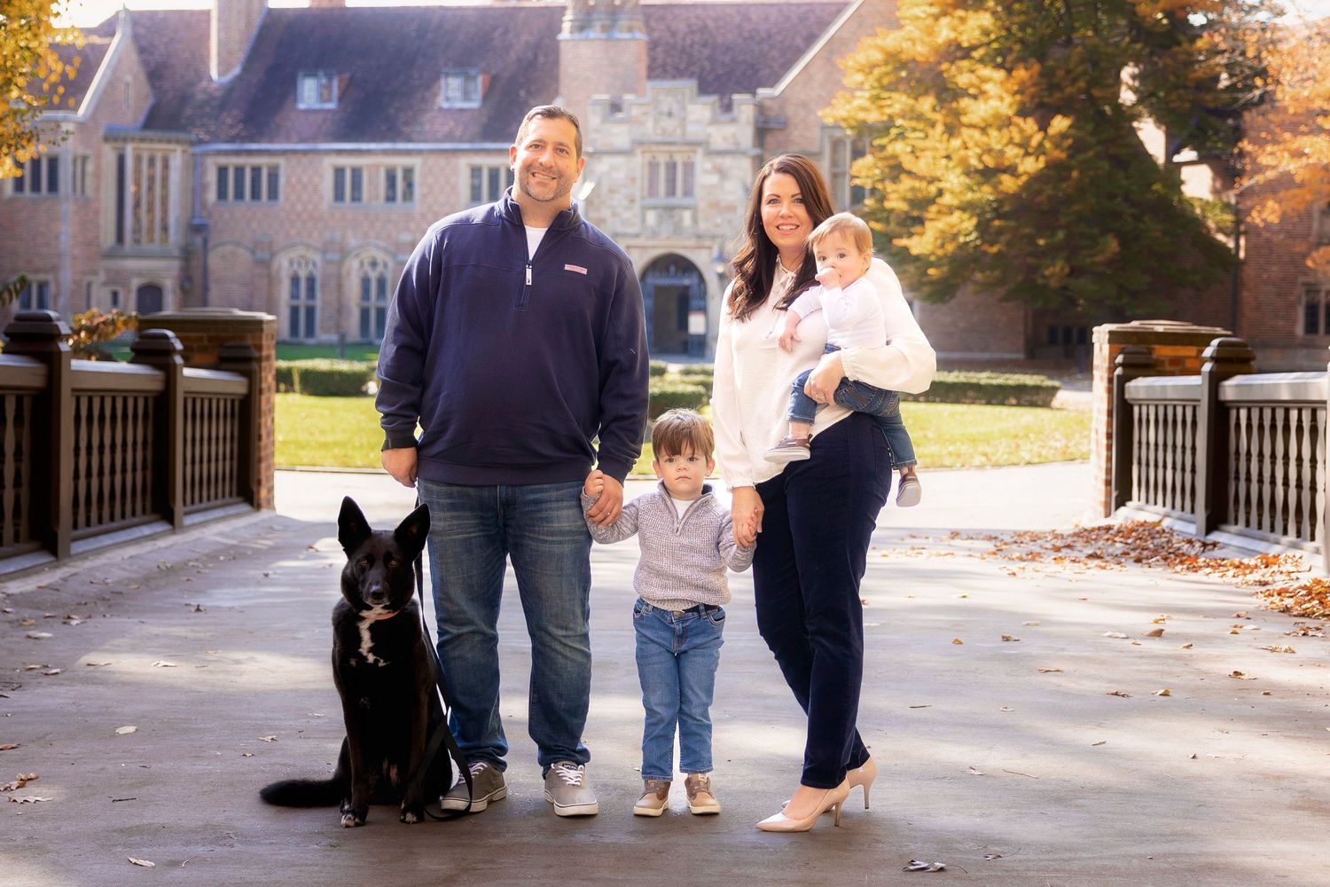 Family with black dog poses together outside stately stone building on autumn day.