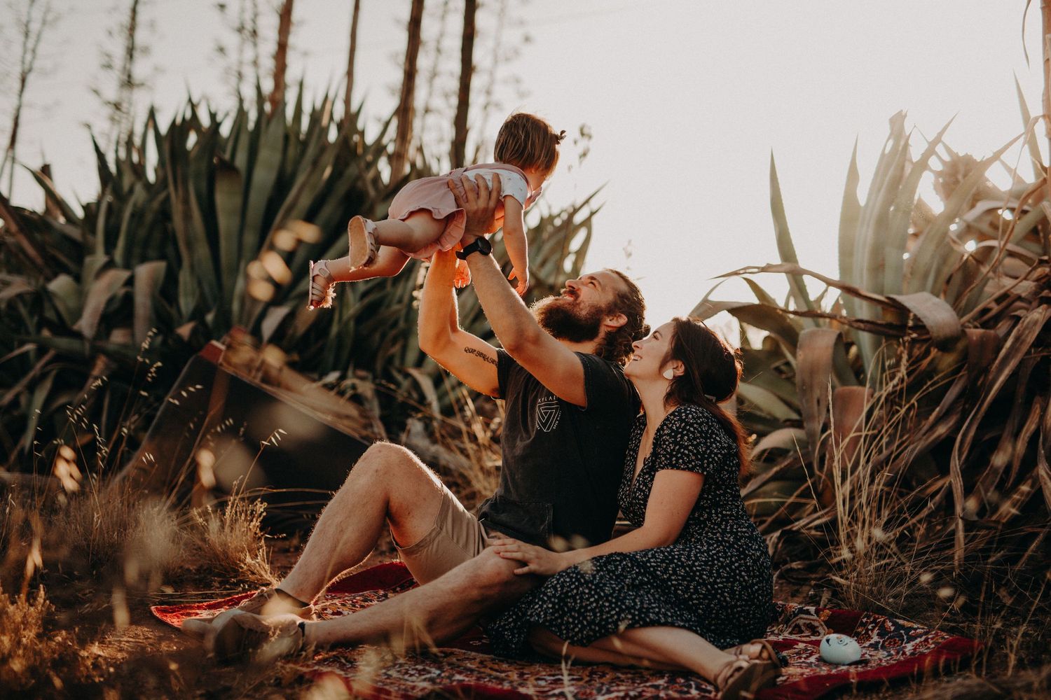 A family relaxes together on a patterned blanket during golden hour in a natural outdoor setting.