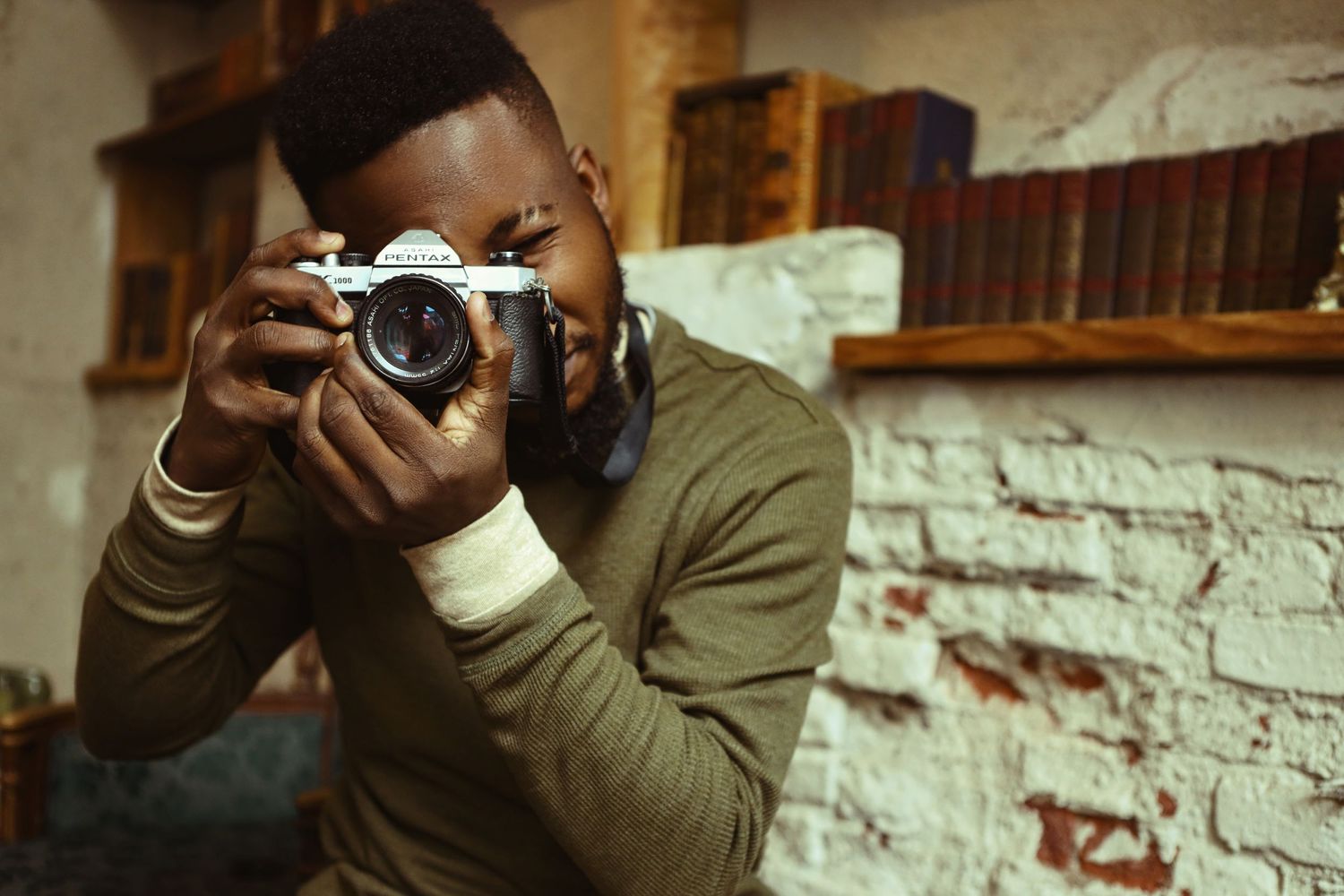 Person in olive sweater photographing with vintage camera against white brick wall.