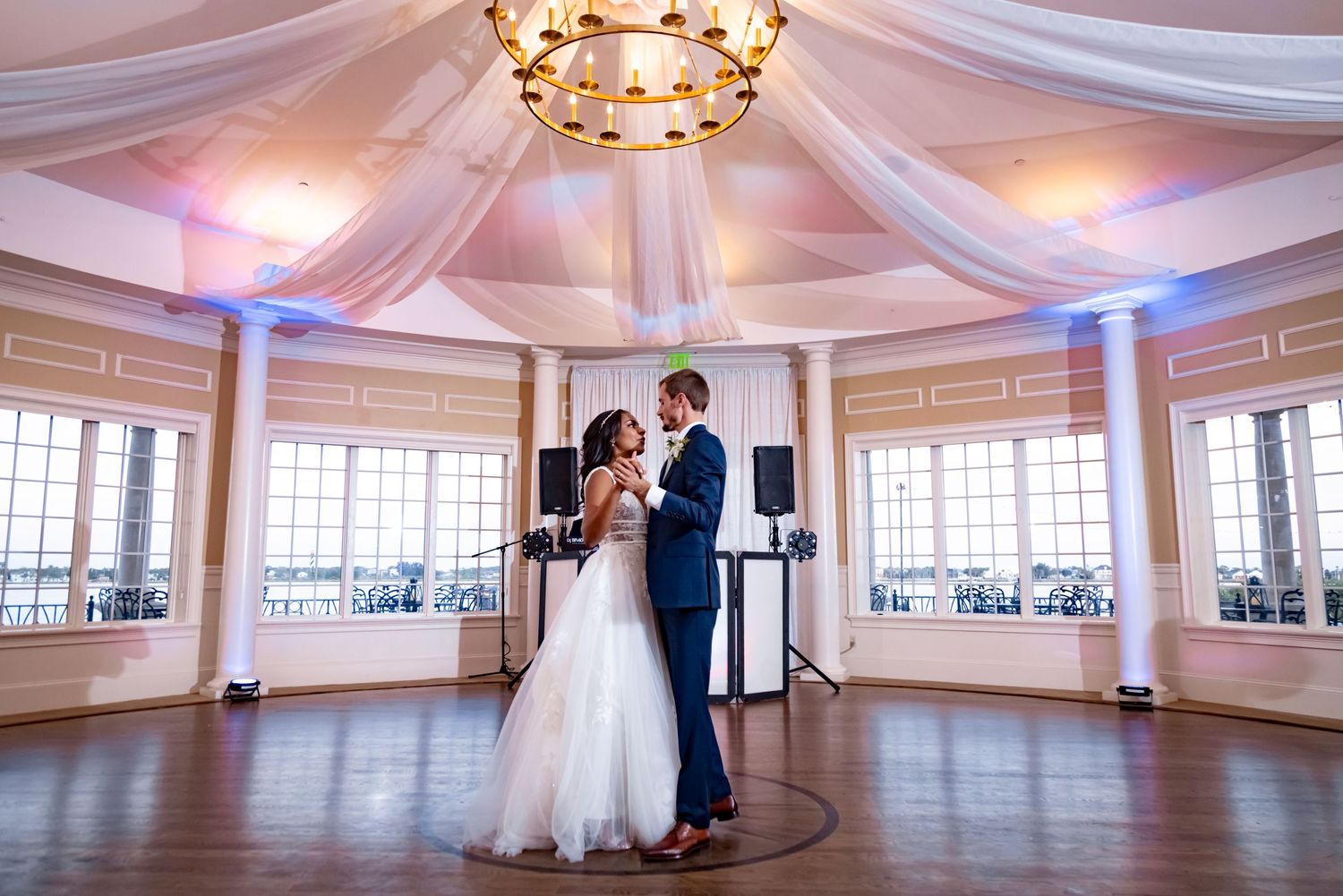 A couple sharing their first dance in an elegant ballroom with draped ceiling and chandelier.