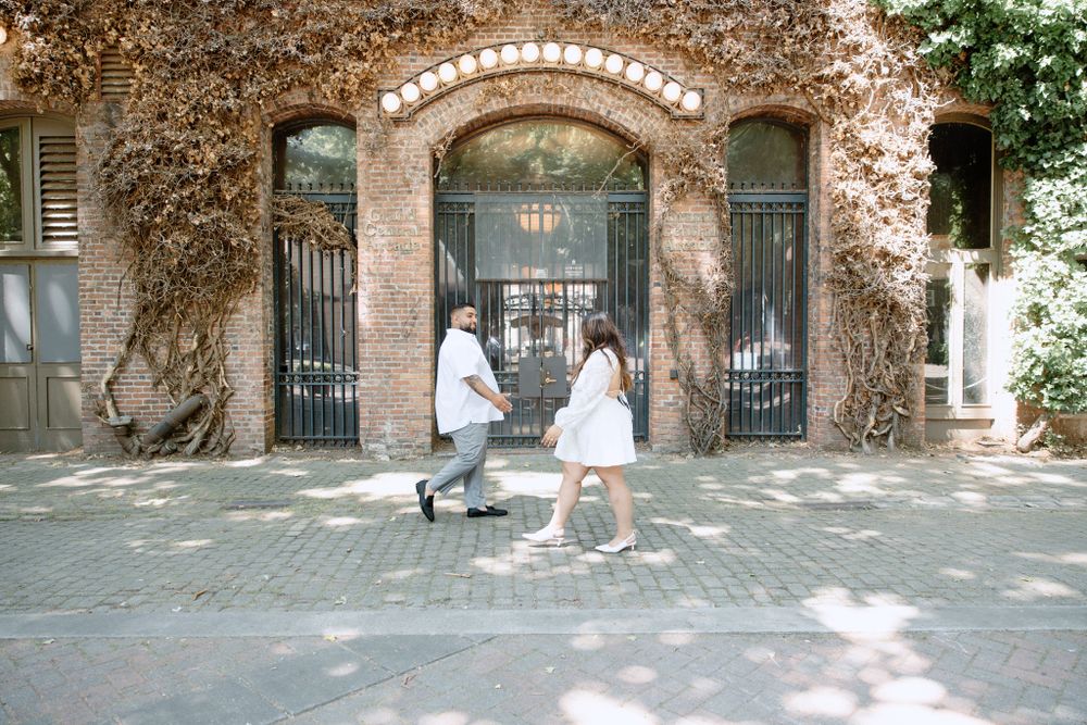 Two figures in white dresses walk through an ivy-covered archway entrance on a sunny urban street.