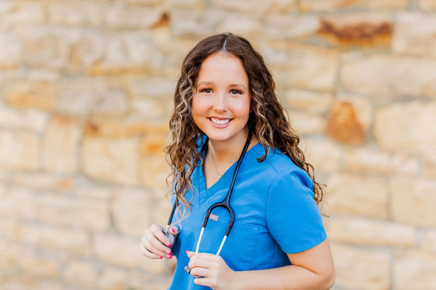 Healthcare professional in light blue scrubs smiling warmly against a stone wall background.