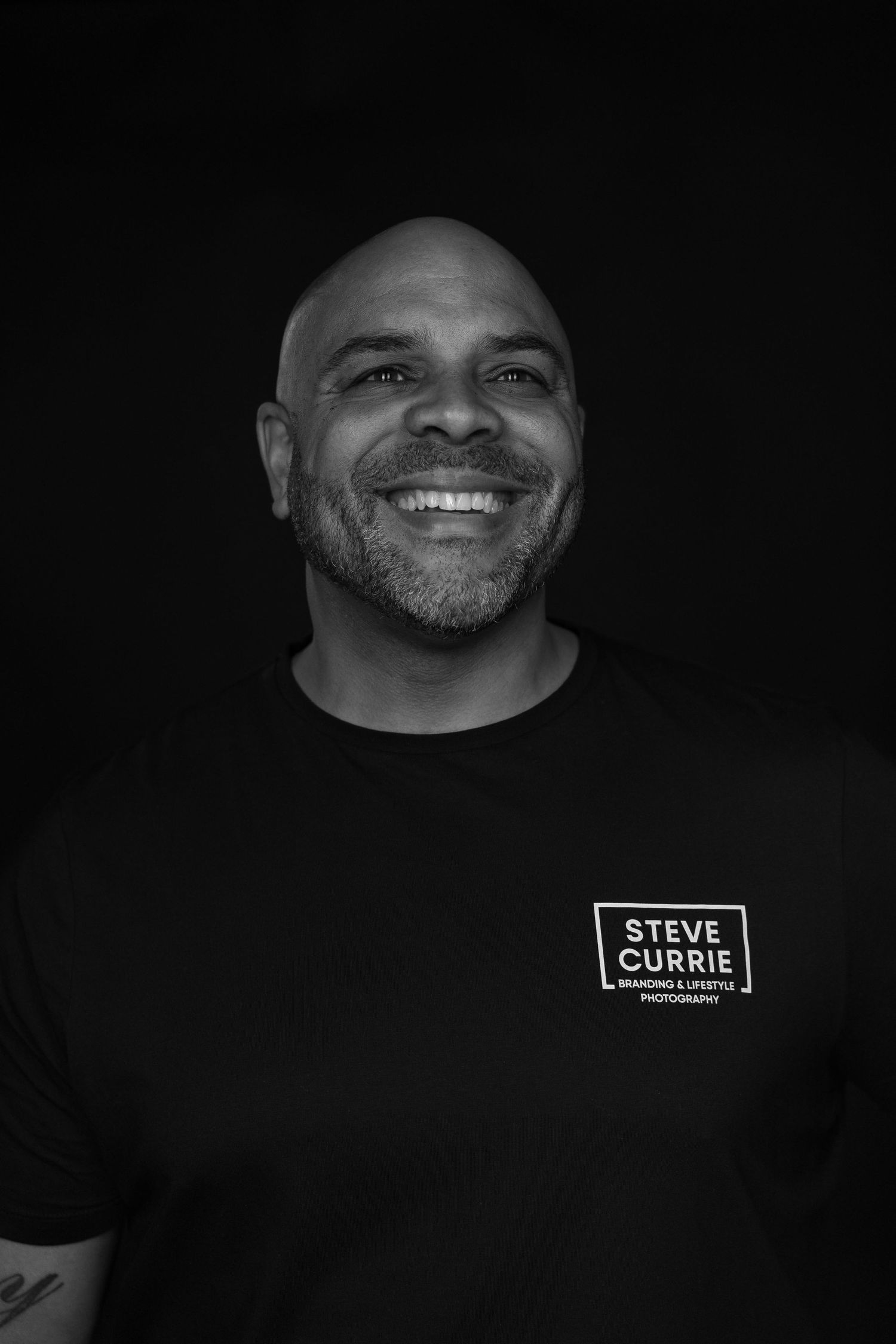 Black and white portrait of smiling man wearing Steve Currie branded t-shirt.