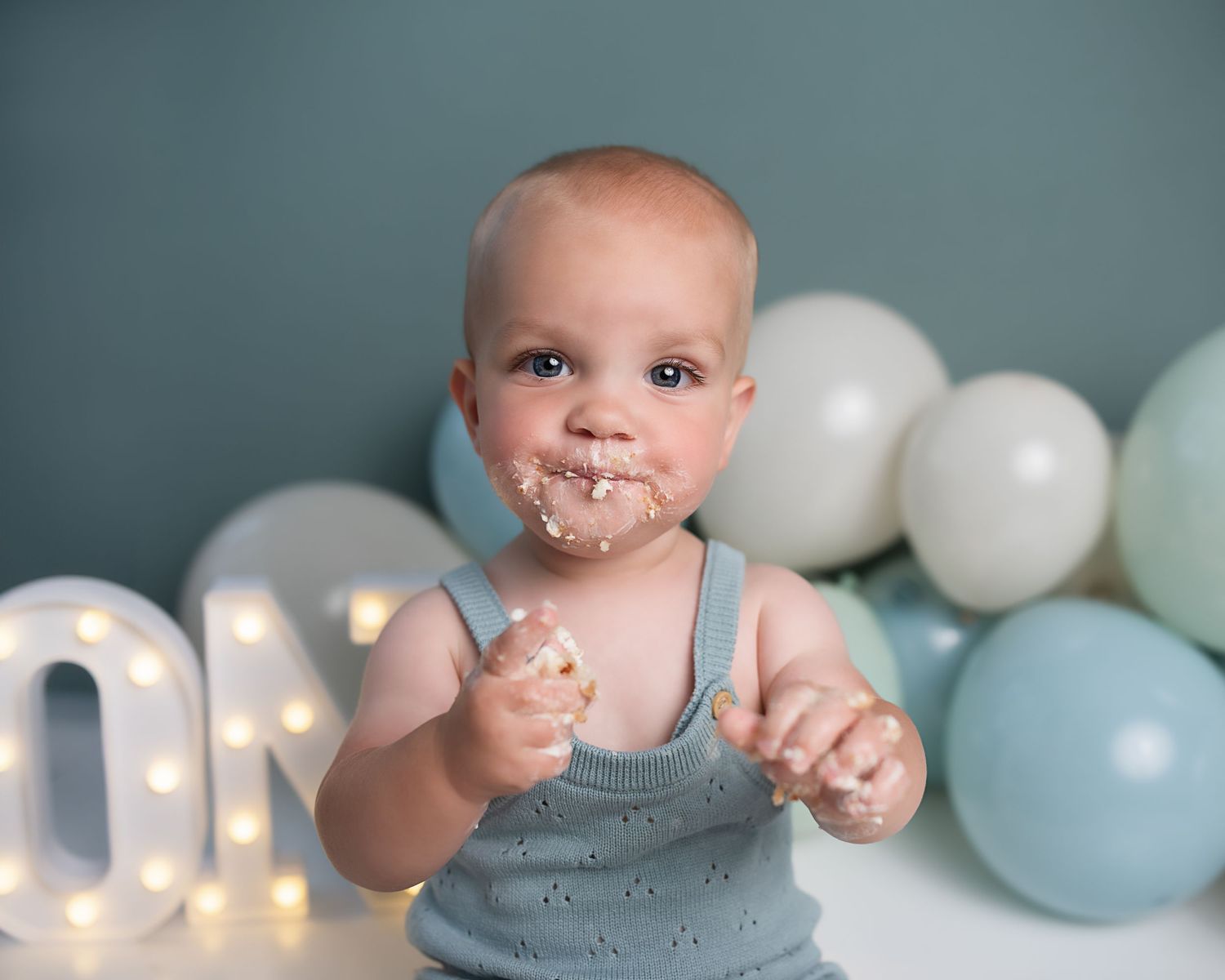 Baby celebrating first birthday smashing cake with frosting on face near ONE letter sign.