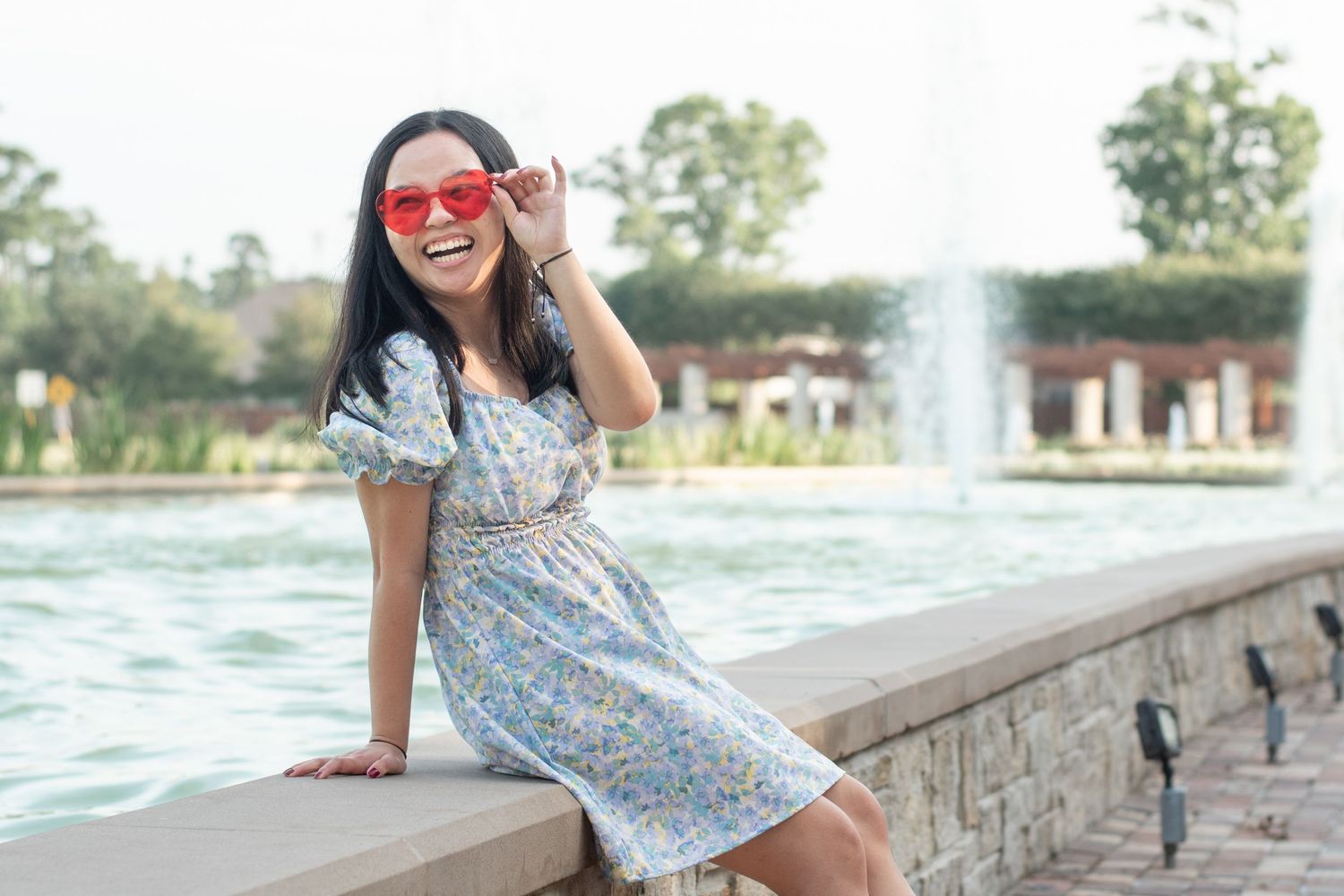 A person in a floral dress and red sunglasses poses by a fountain in a sunny park setting.