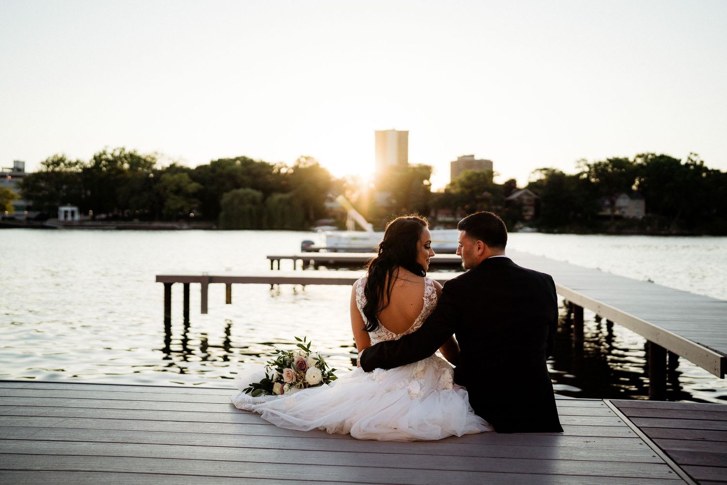 Couple sitting on wooden dock overlooking lake at sunset in formal attire.