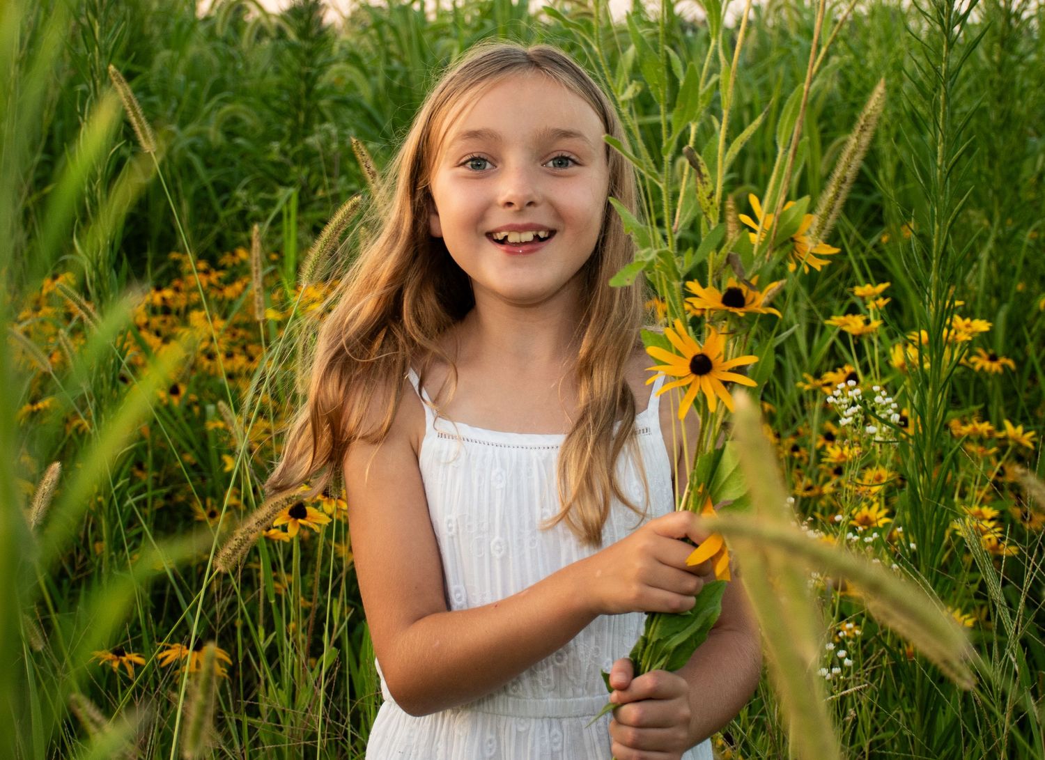 A young girl in a white dress poses with sunflowers in a lush summer garden.