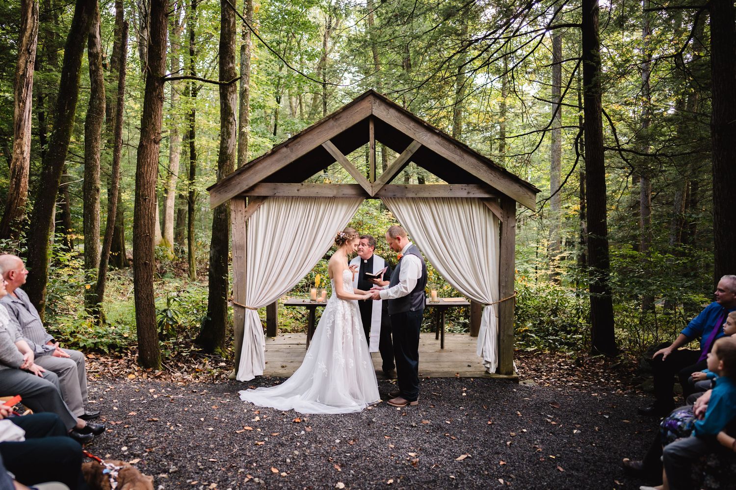 Outdoor wedding ceremony setup with wooden gazebo and chairs in a wooded forest setting.