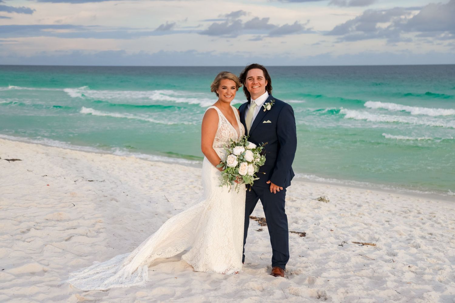 Romantic beach wedding portrait with turquoise ocean waves and cloudy sky backdrop.