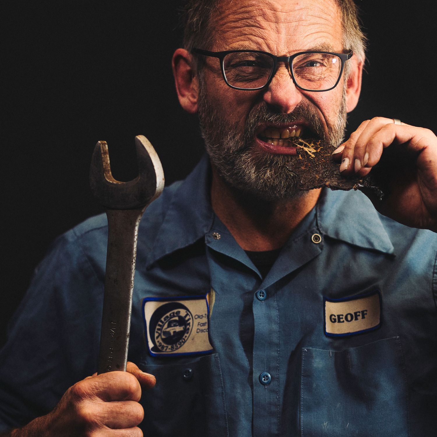 A mechanic in work uniform holds a wrench while posing against a dark background.
