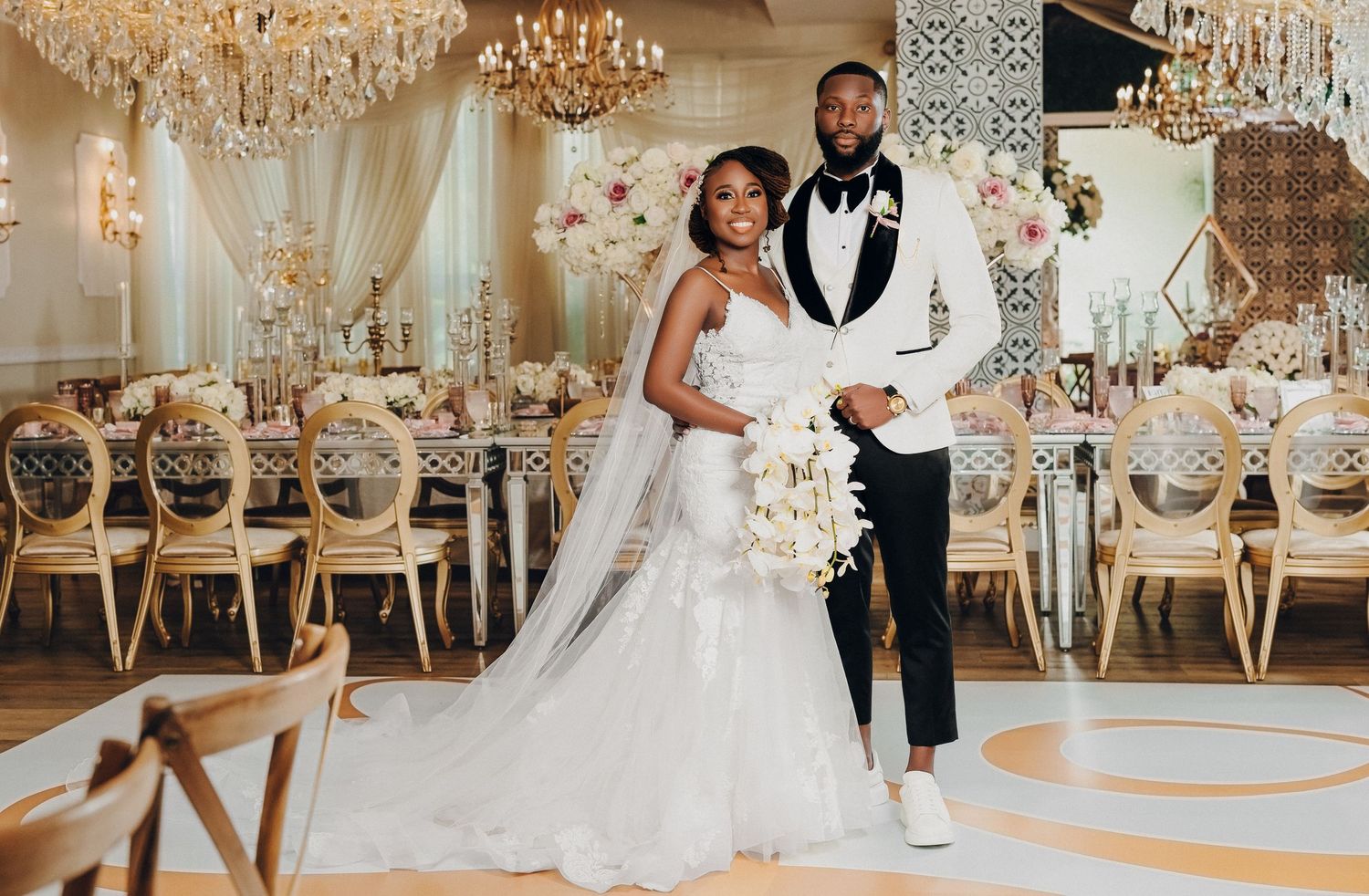 Black bride and groom pose in opulent gold ballroom with chandeliers, floral arrangements, and white dance floor.